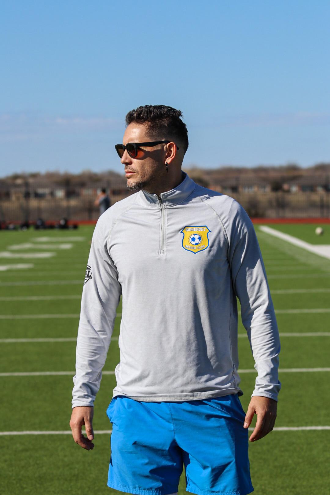 Man wearing sunglasses and a light gray jacket with a logo, standing on a football field with a blue sky.
