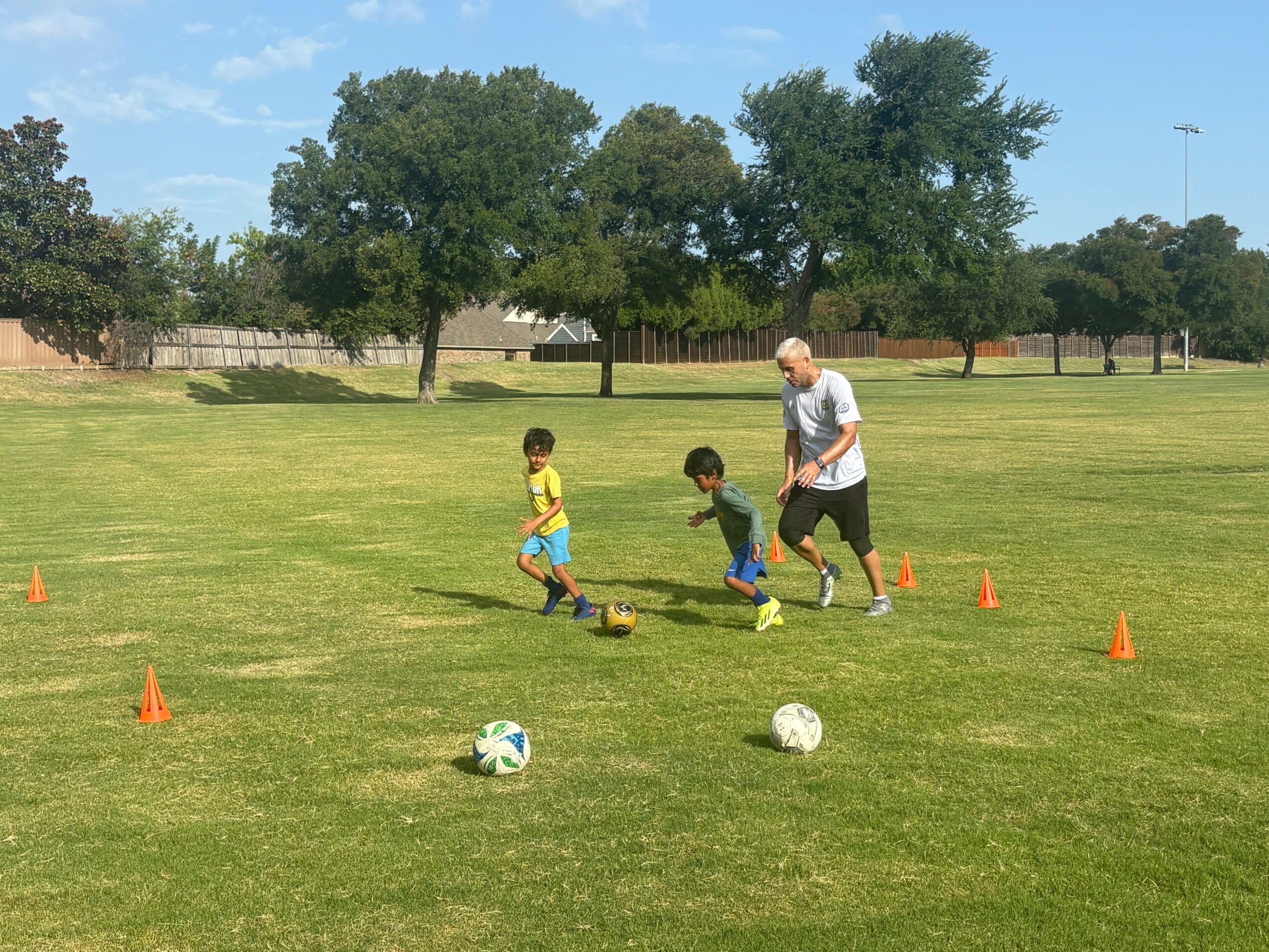 Two children and an adult playing soccer on a grassy field with orange cones, trees, and a fence in the background.