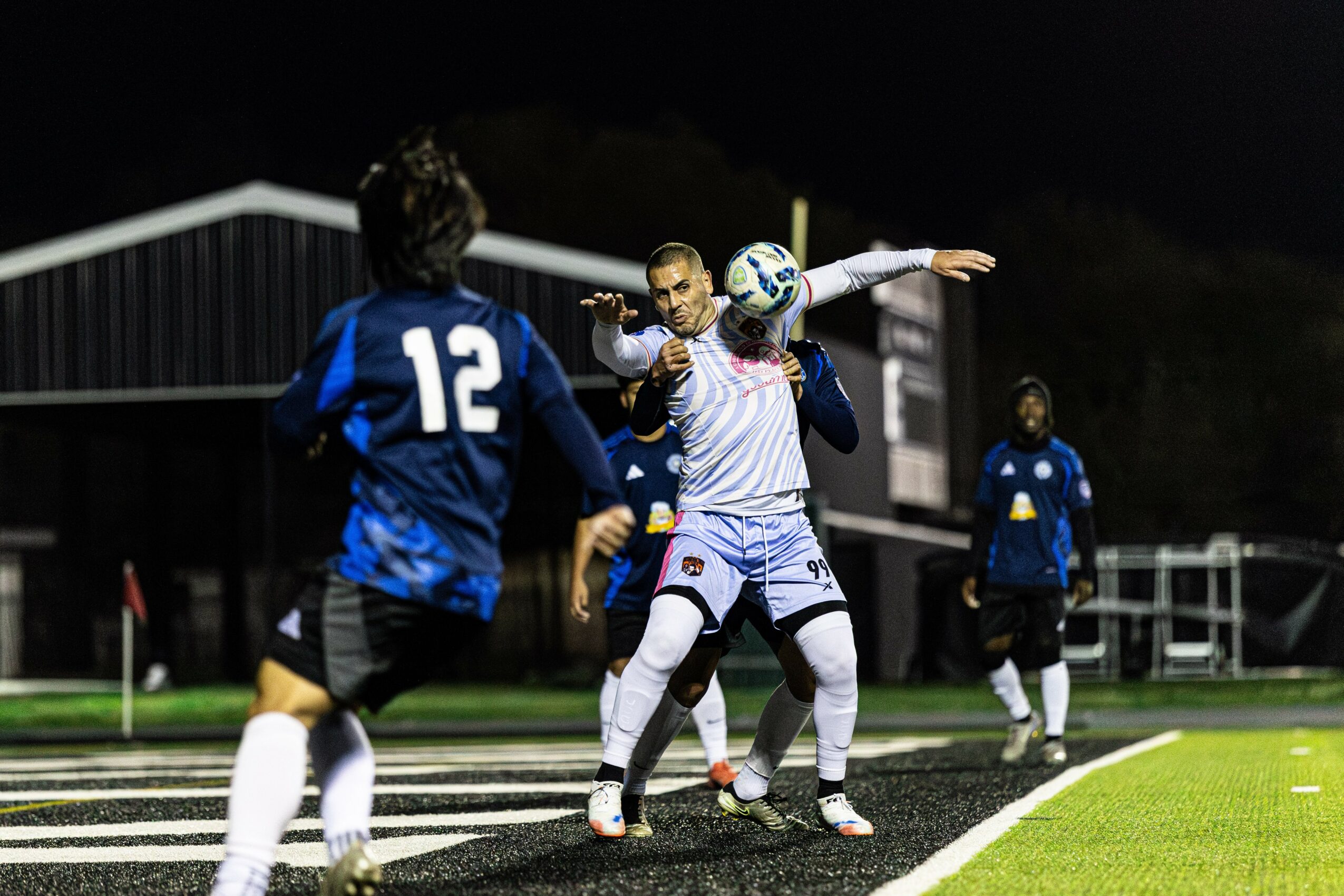 Soccer players in action on the field at night, with one player jumping to head the ball, others watching.