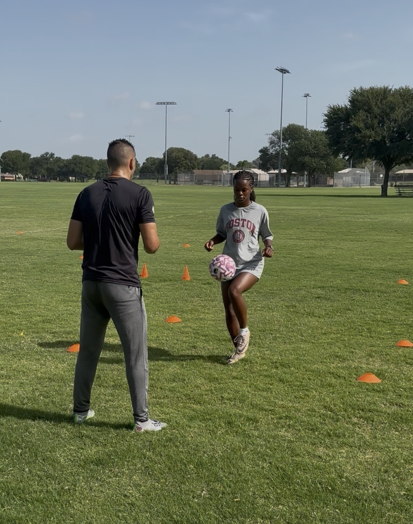 Person holding a soccer ball in a grassy field with another person facing them, orange cones set up for training, trees and light poles in background.