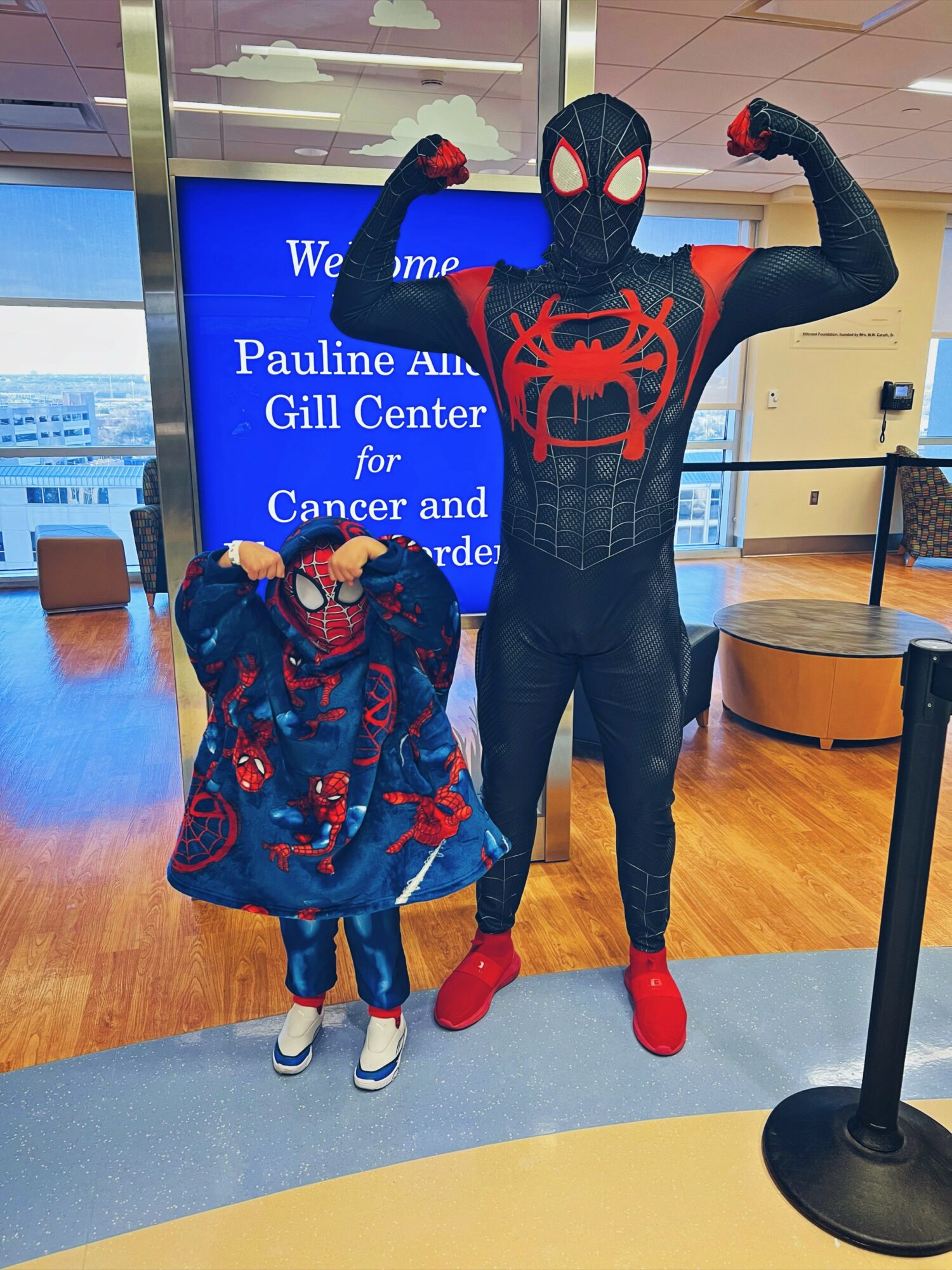 Person dressed as Spider-Man and a child in a Spider-Man costume pose indoors near a welcome sign, with a blue background and wooden floor.