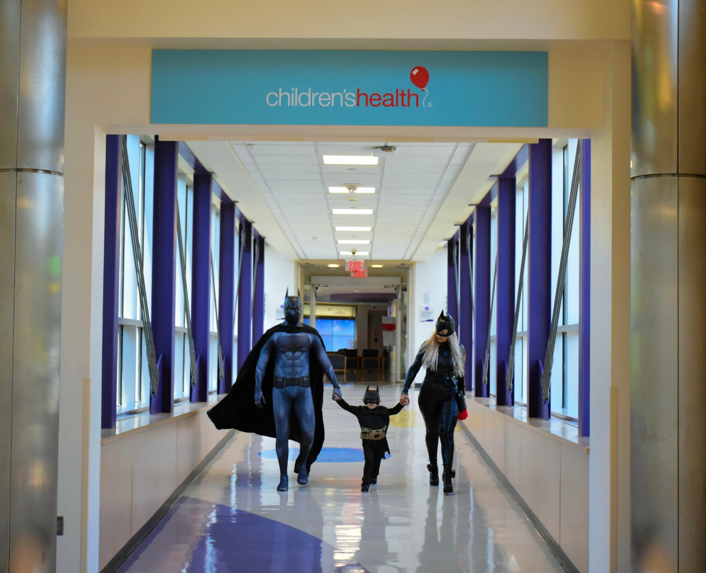 A child holding hands with two adults walking through a hospital corridor with blue curtains and a sign reading 'children's health'.