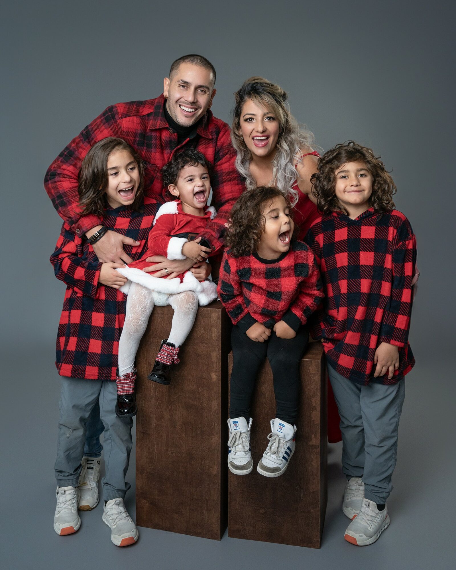 Family of six wearing matching red and black plaid outfits, smiling together against a gray background.