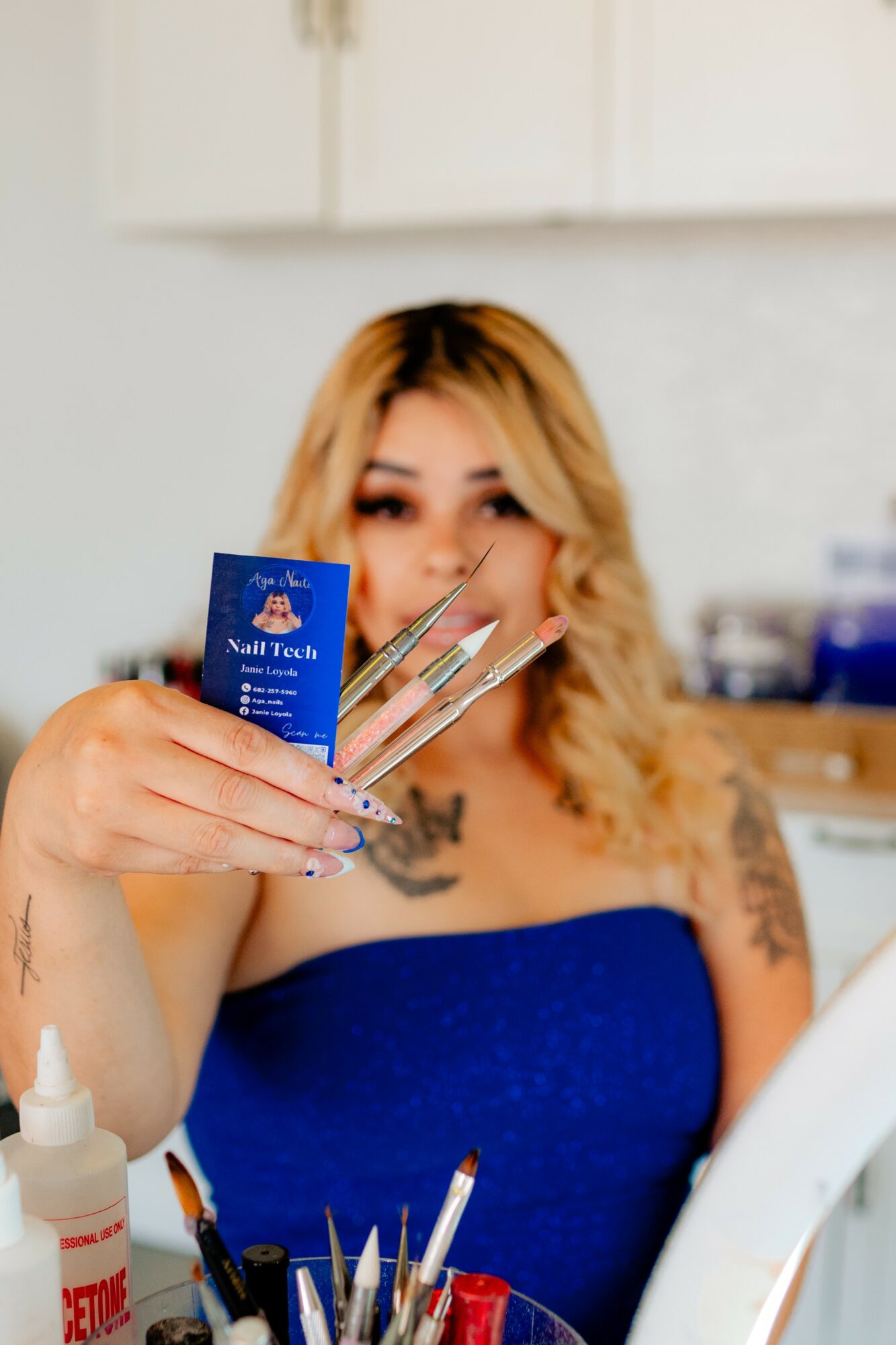 Woman with blonde hair holding nail files and a blue nail polish card, smiling in a salon setting.