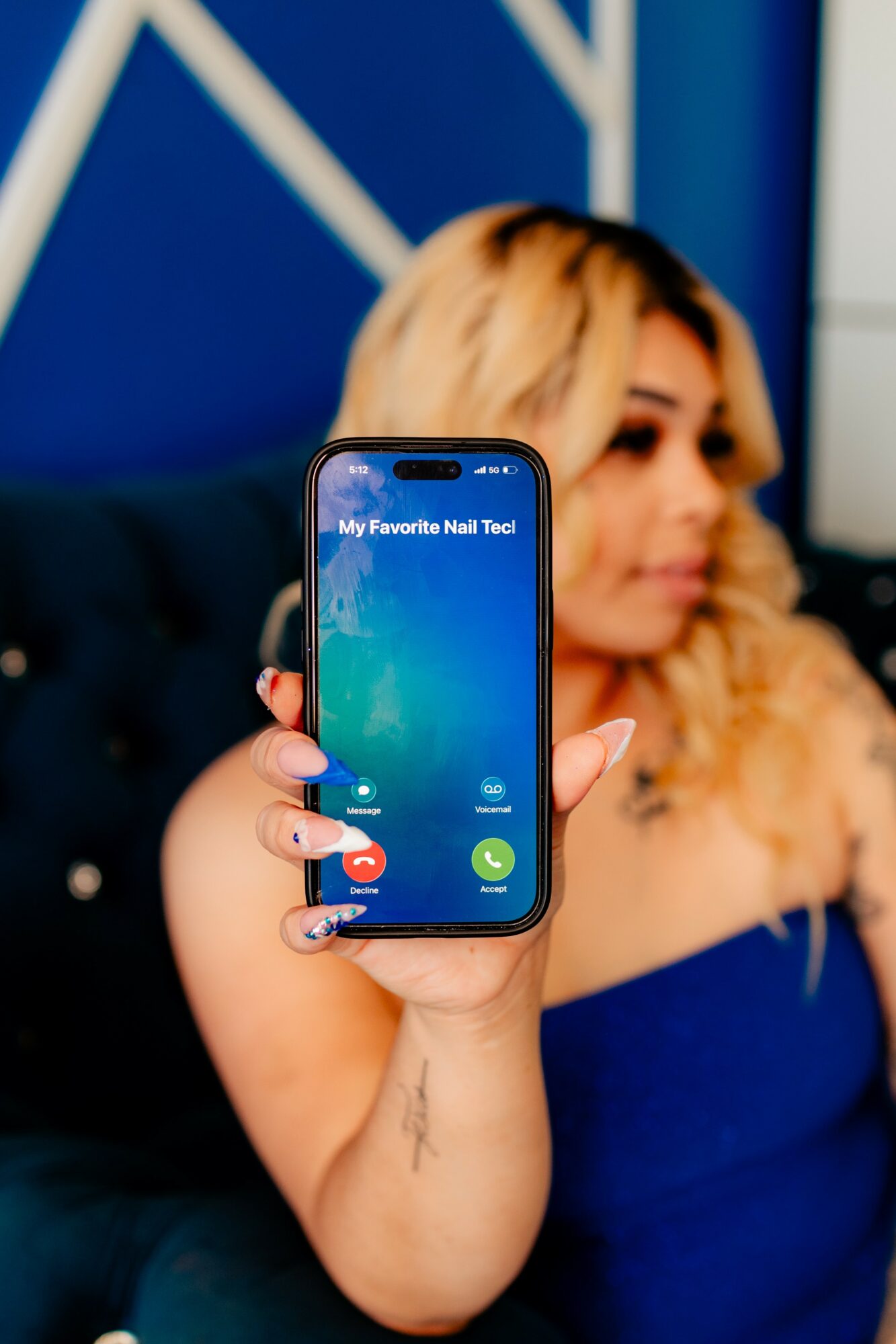 Person with blonde hair holding a smartphone displaying a nail tool app, in a room with blue walls and furniture.