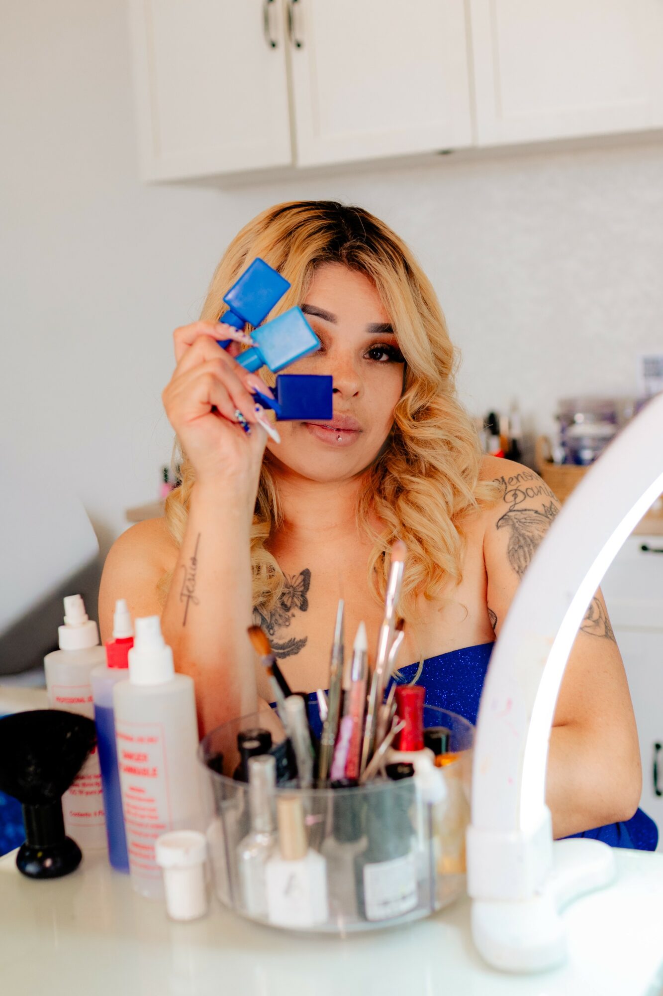 Woman with blonde hair holding blue makeup sponges, surrounded by makeup products and brushes, in a well-lit room.