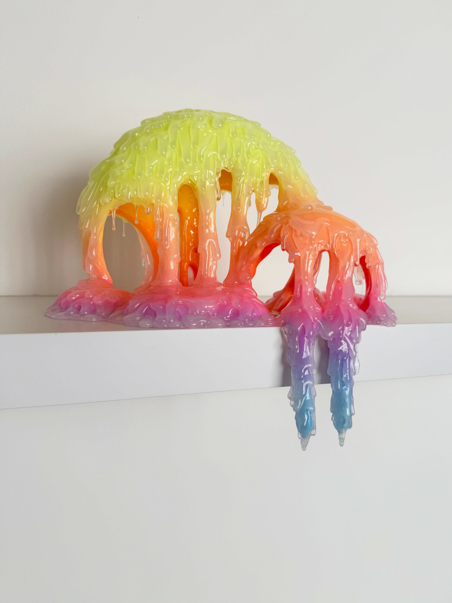 Colorful melting wax sculpture with drips, in yellow, orange, pink, purple, and blue, on a white surface.
