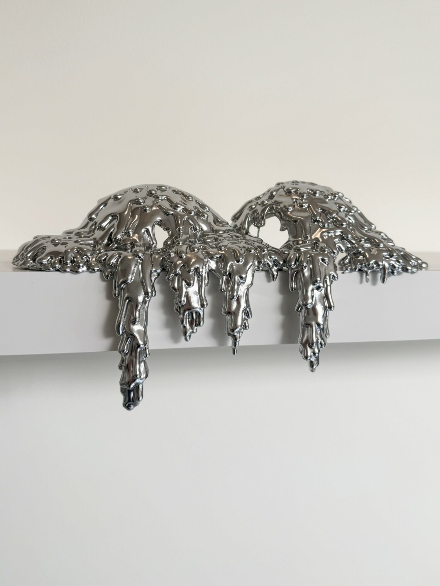 Metallic sculpture with dripped, melting appearance on a white surface against a plain background.