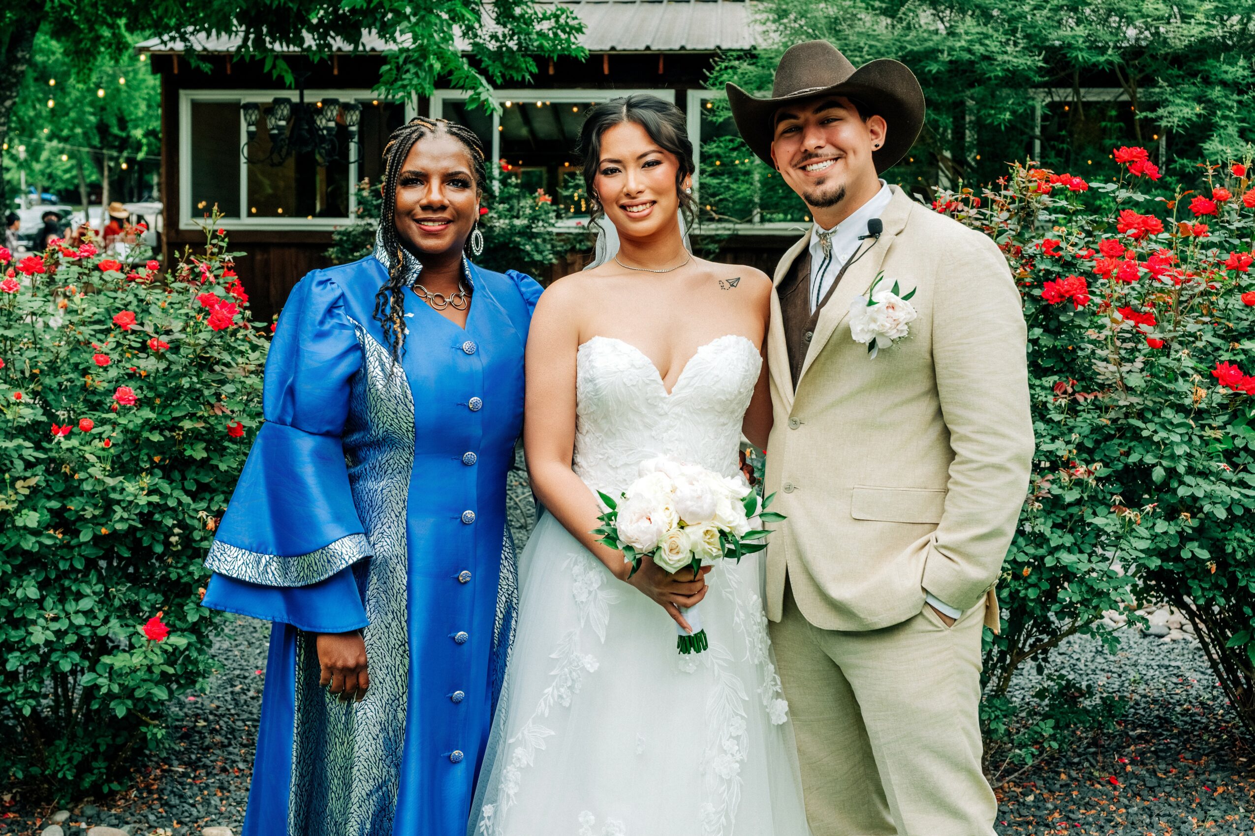 Three people in wedding attire standing outdoors with red and pink flowers and greenery in background.