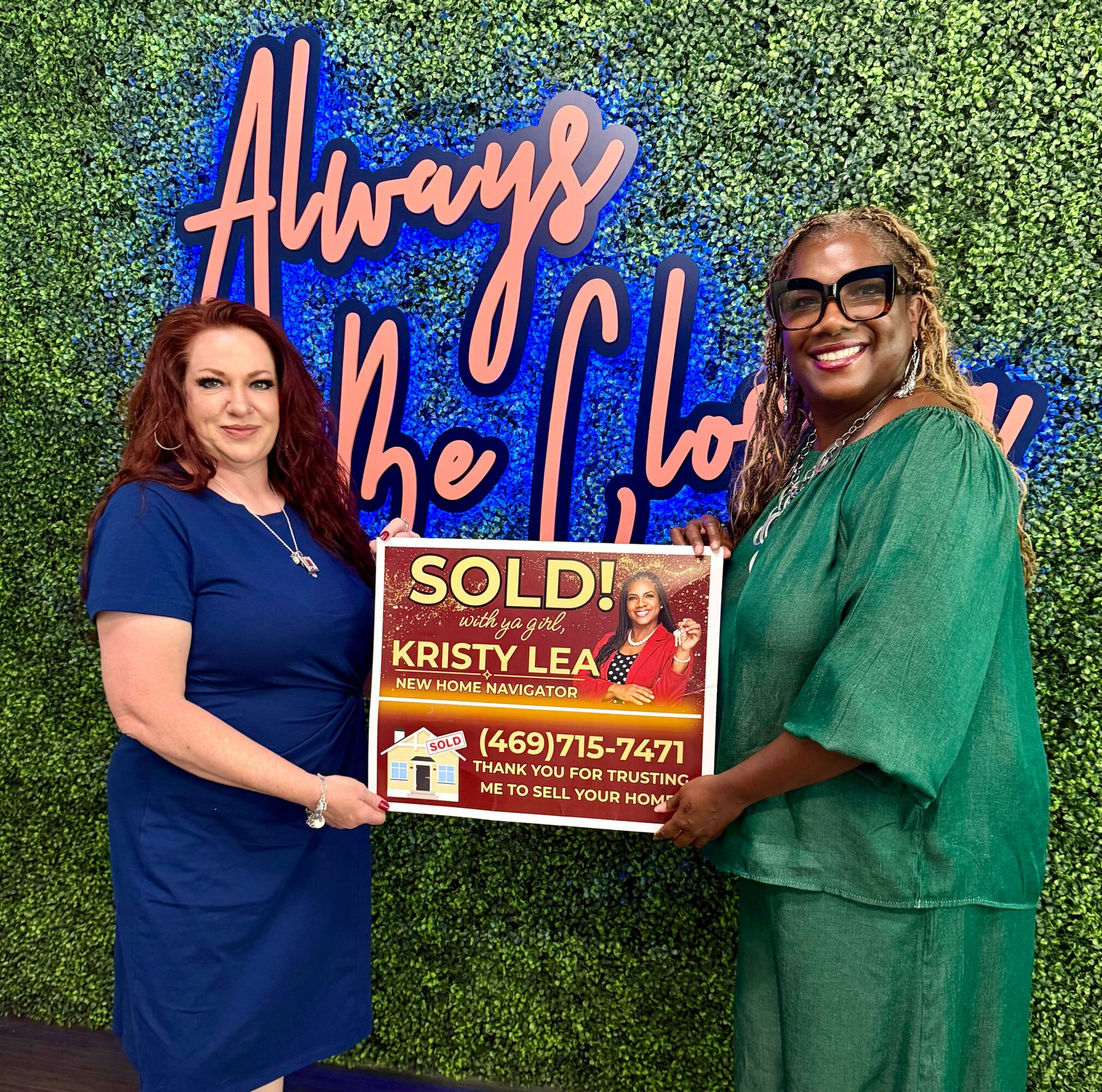 Two women standing in front of a green wall with neon sign, holding a sold sign for a house, smiling.