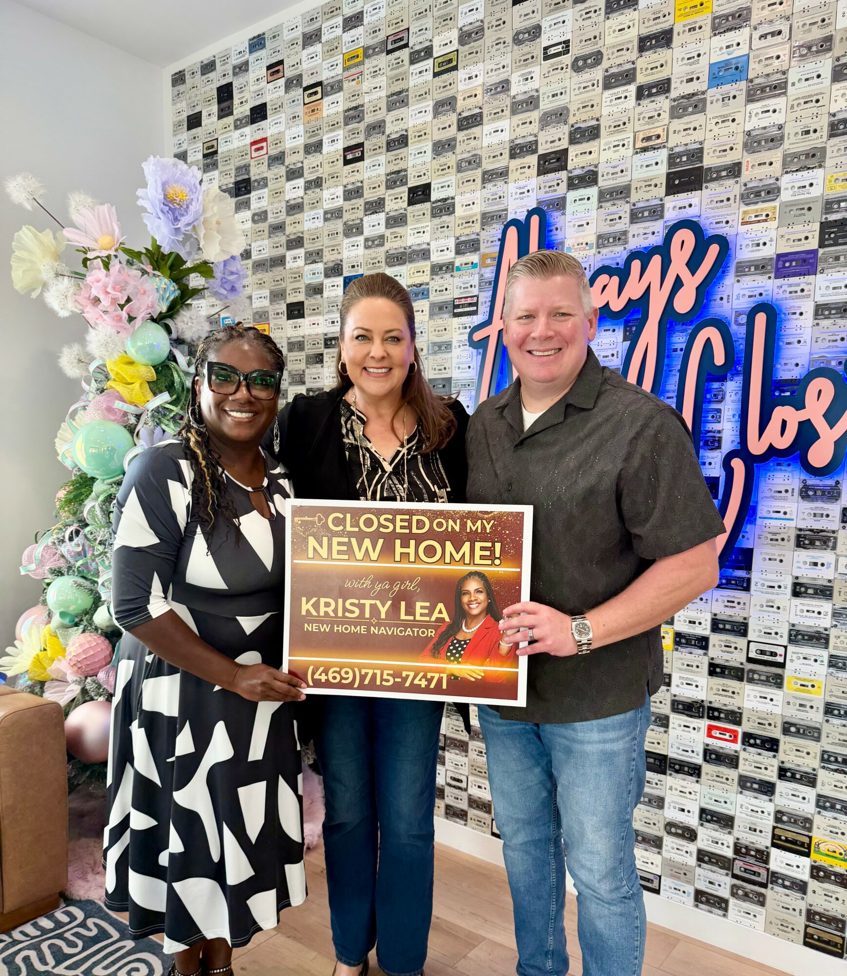 Three people smiling, holding a sign about a new home, standing in front of a wall with a neon sign and flowers.