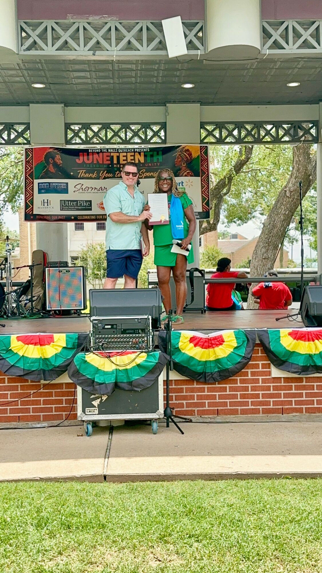 Three people on stage holding awards, with a banner and colorful decorations behind them, outdoors during daytime.