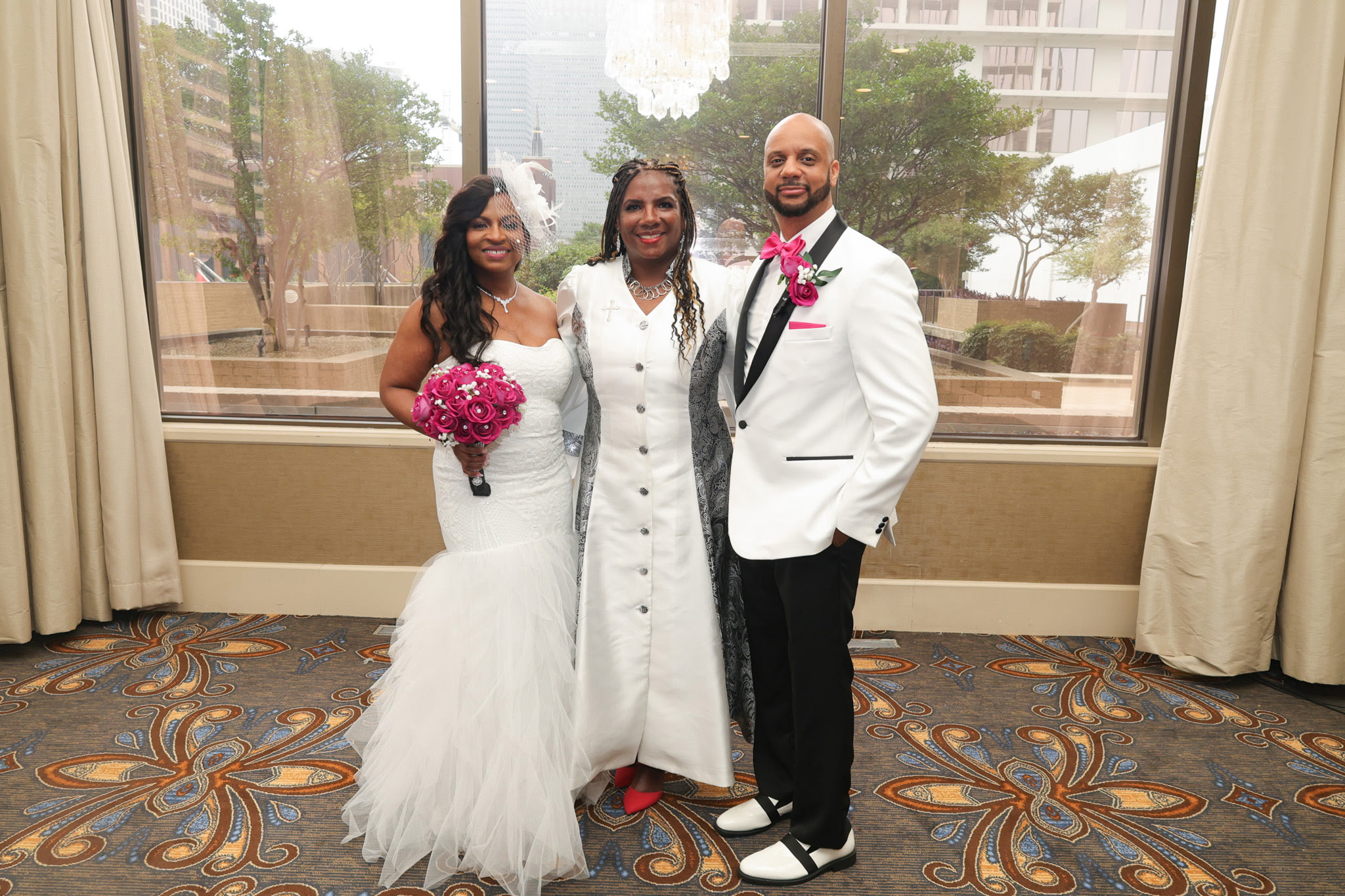 Three people in wedding attire standing indoors in front of large windows, smiling, with a patterned carpet below.