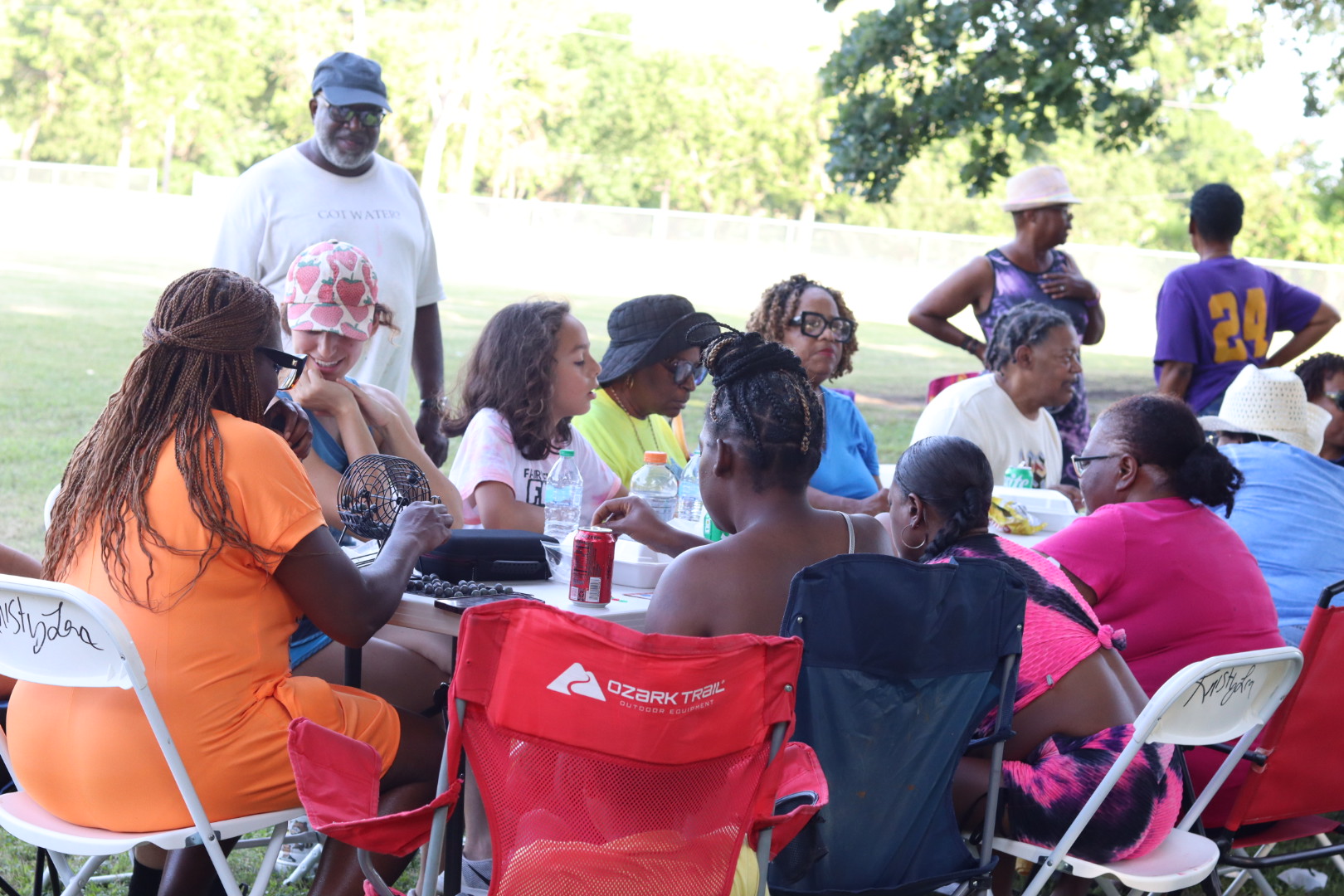 Group of people sitting at a table outdoors, some standing, with trees and grass in the background.