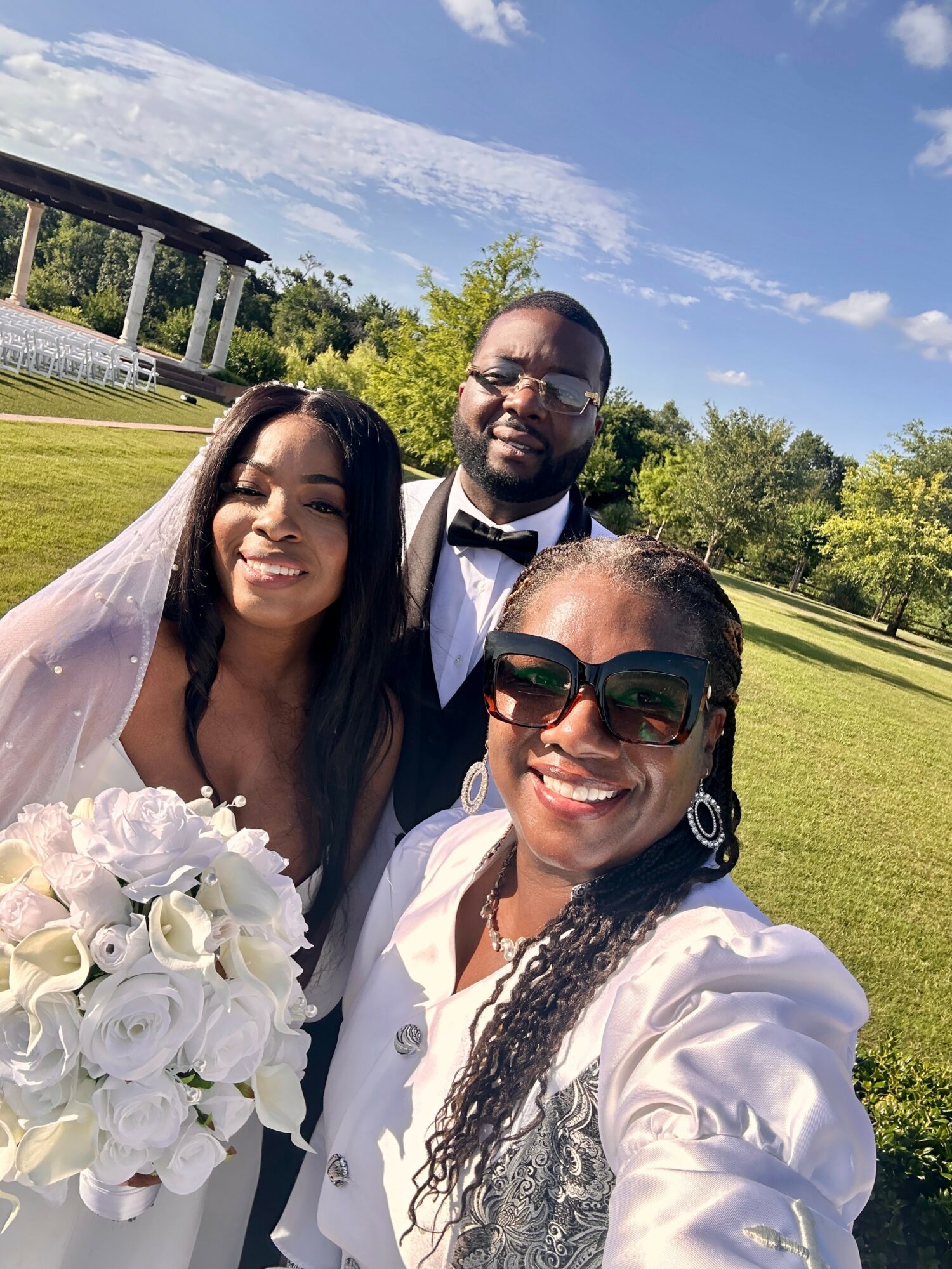Three people smiling outdoors on a sunny day, with a woman holding a bouquet of white flowers, all dressed formally.