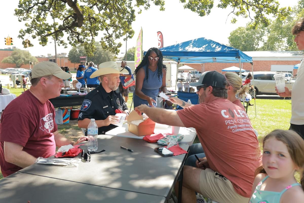People sitting at a table outdoors, with a woman standing nearby, under a blue canopy, trees, and parked cars in background.