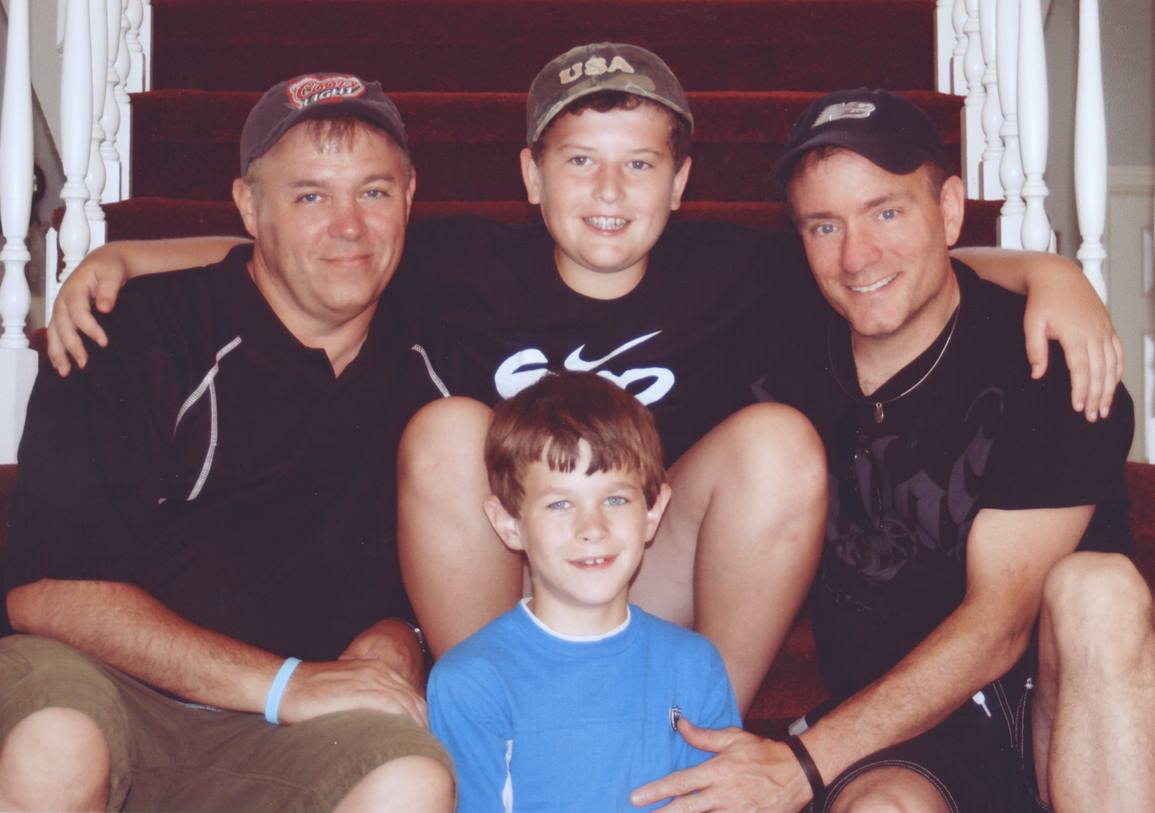 Three men and two boys sitting on stairs, smiling, with arms around each other, indoors. One boy is sitting on a man's lap.