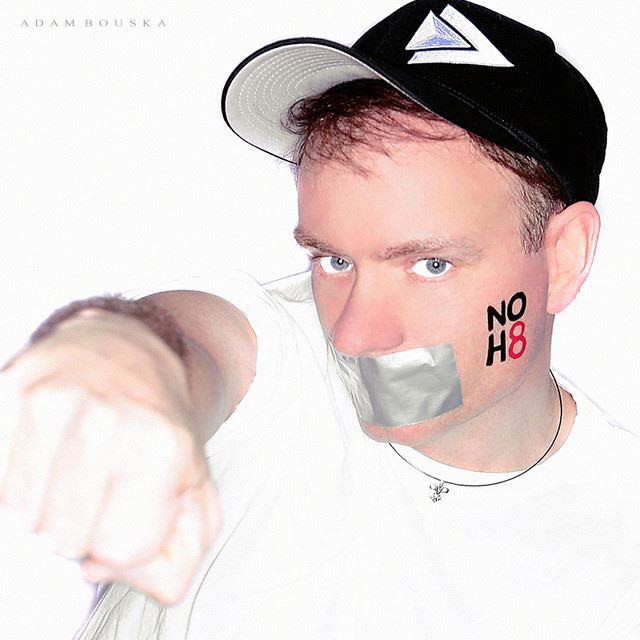 Person with tape over mouth, wearing a cap, pointing forward, with 'NO H8' on cheek, against a white background.