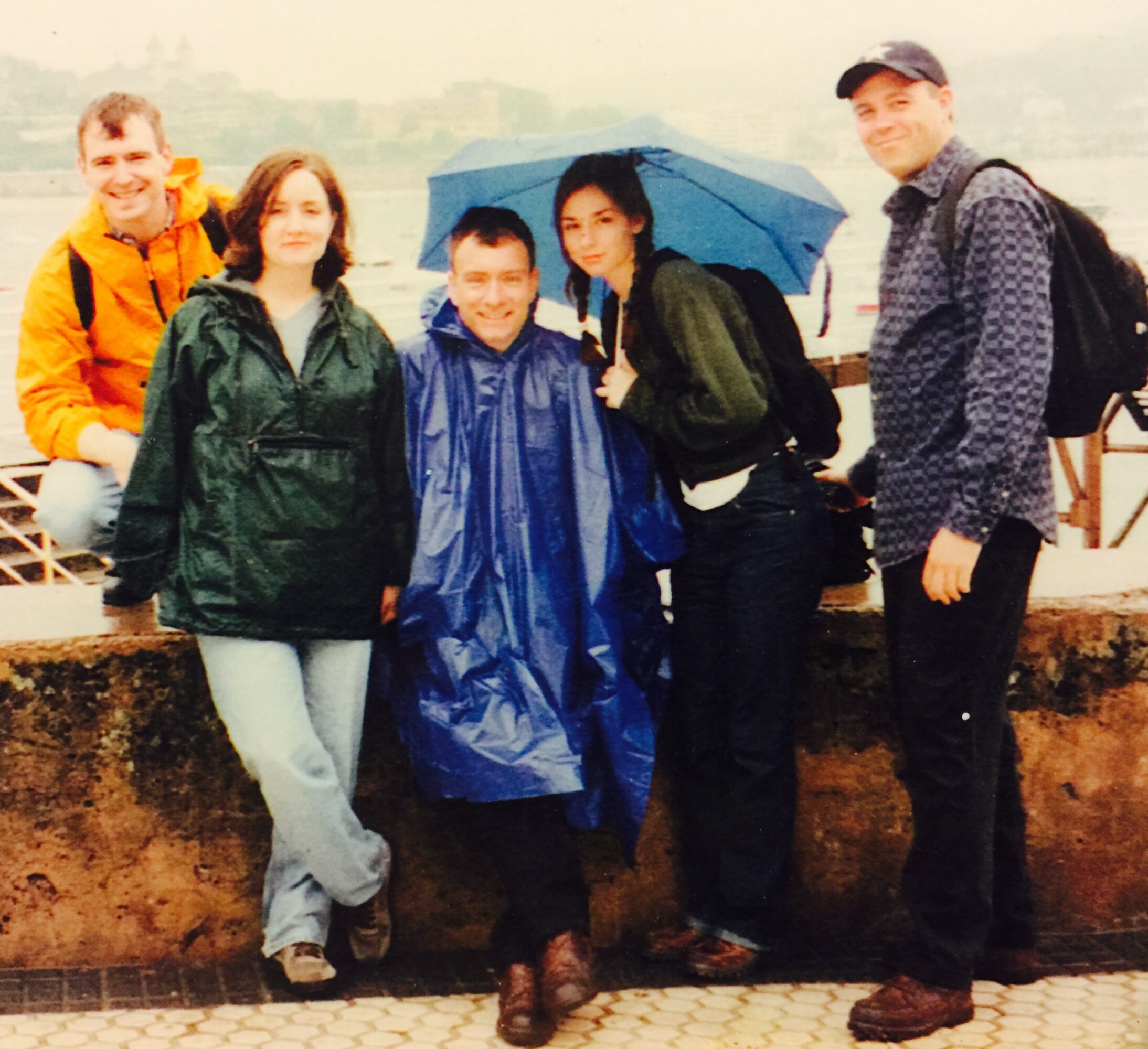Group of five people standing outdoors in rainy weather, some holding umbrellas, smiling, with a body of water in the background.
