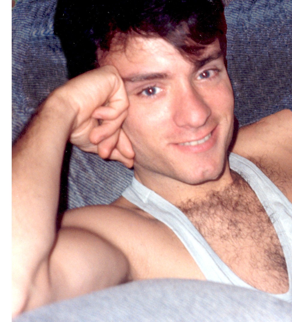 Young man lying on a couch, smiling, with his head resting on his hand, wearing a tank top.