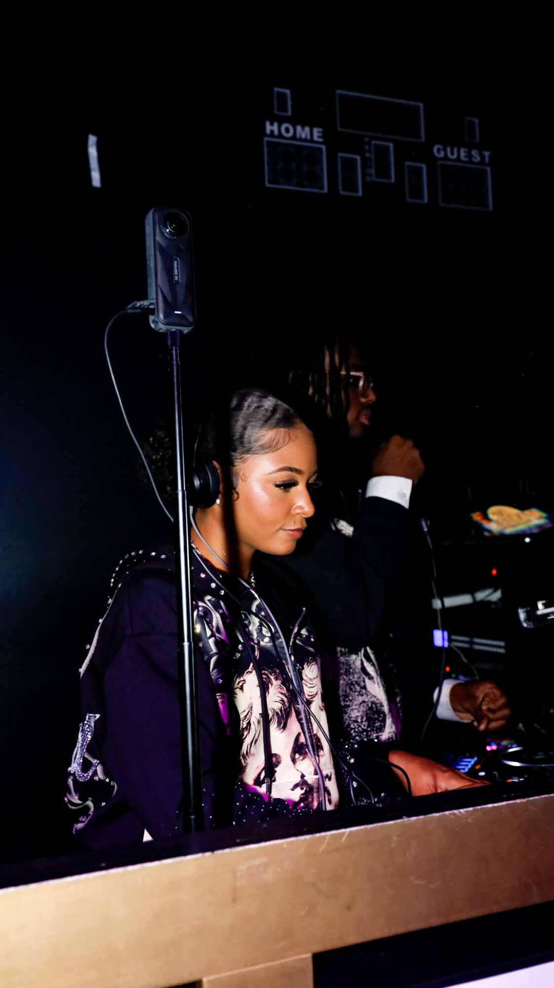 A woman with long dark hair and makeup stands behind a DJ booth, with a person in the background, in a dark setting.