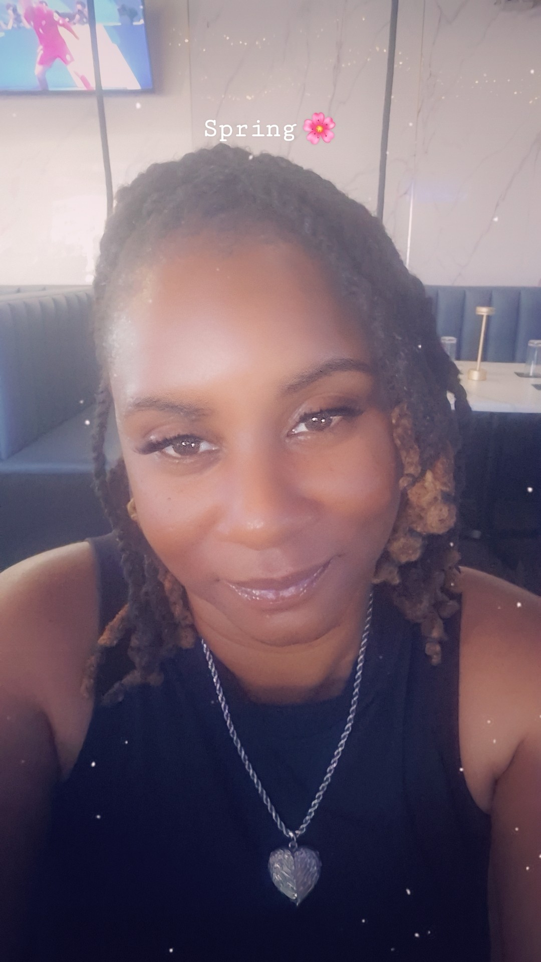 Close-up of a smiling woman with braided hair wearing a necklace with a heart pendant, indoors.