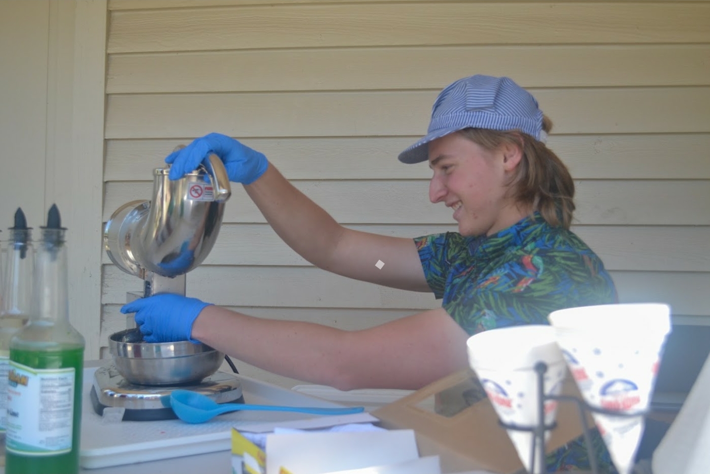 Girl pouring liquid from a teapot into a bowl, wearing a cap and blue gloves, smiling, with a table and cups nearby.