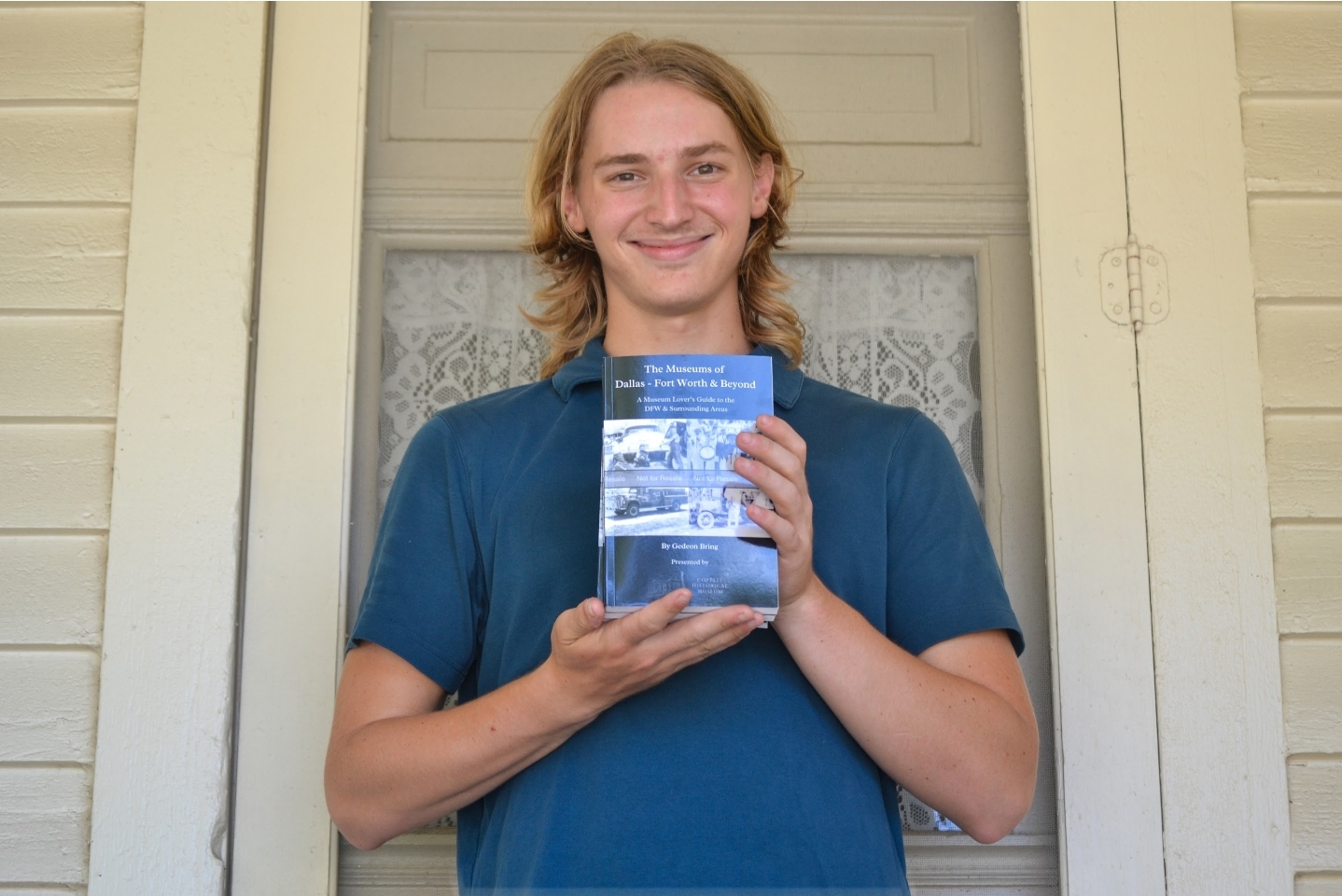 Person with shoulder-length hair smiling and holding a book in front of a door with lace curtains.
