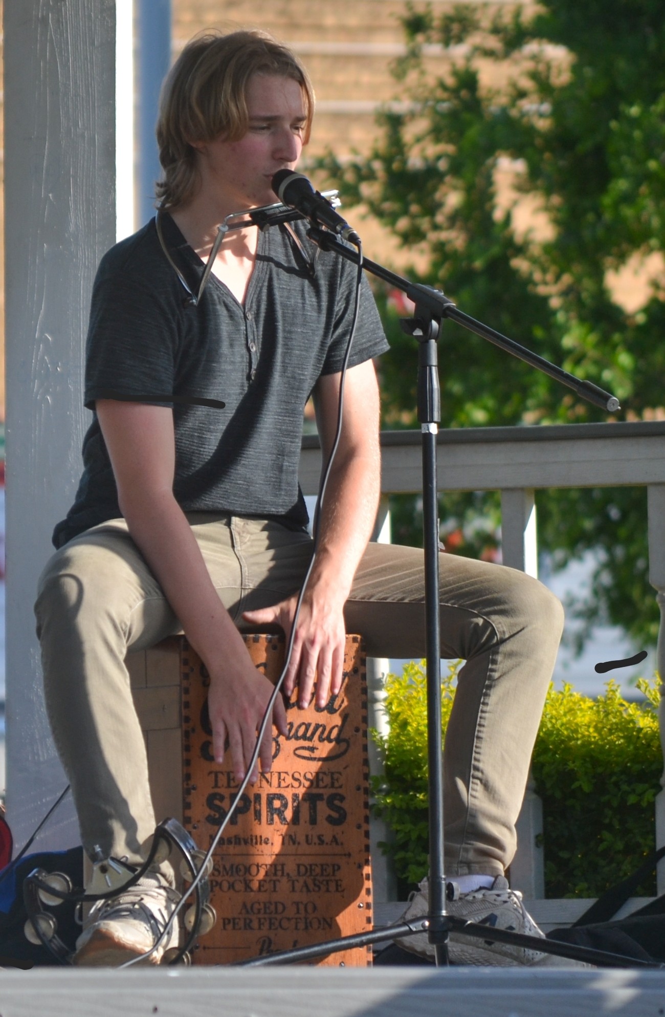 Young person playing a cajón drum and singing into a microphone outdoors, with trees and a building in background.