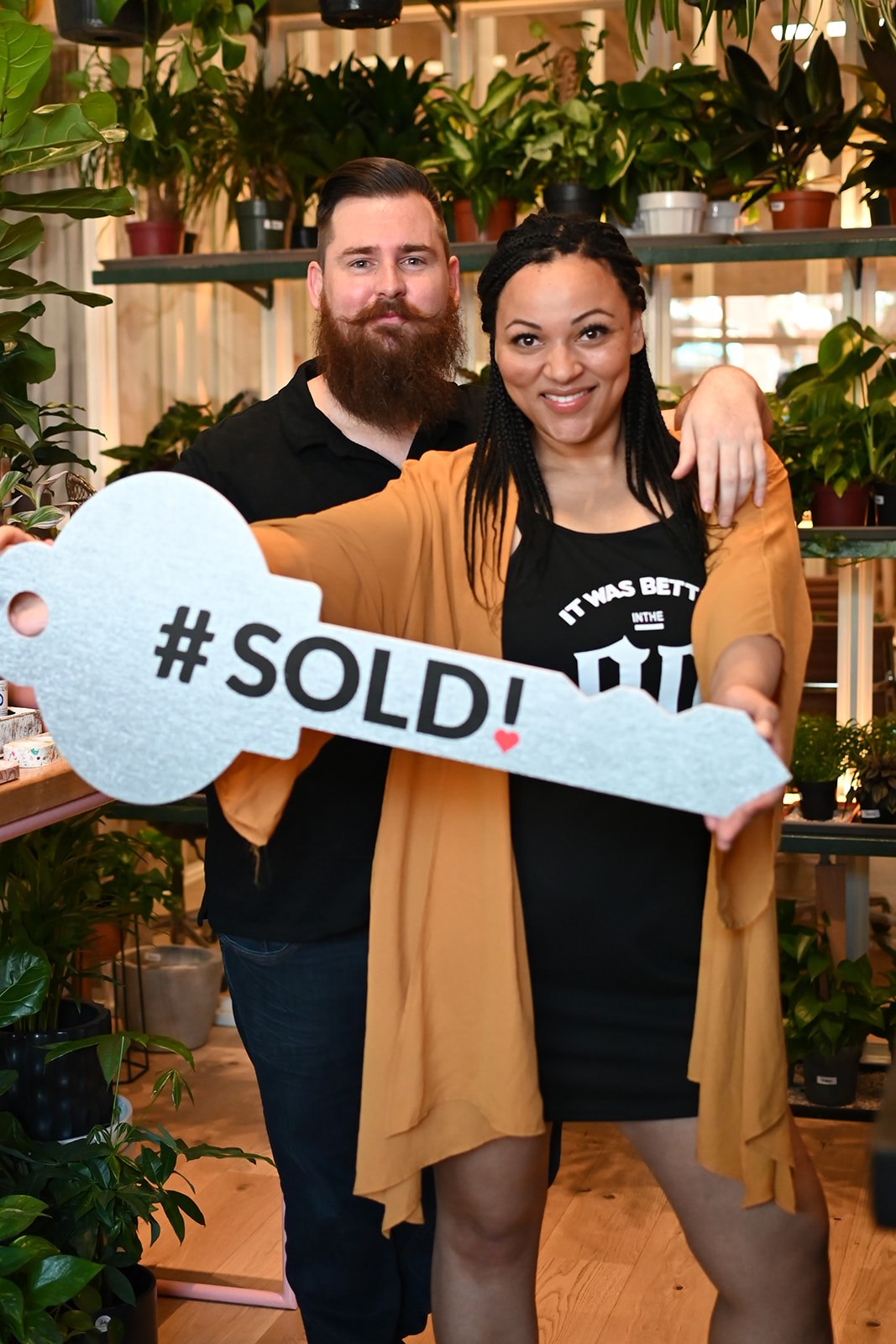 Two people smiling, holding a large key-shaped sign that says '#SOLD!', surrounded by plants in a store or greenhouse.