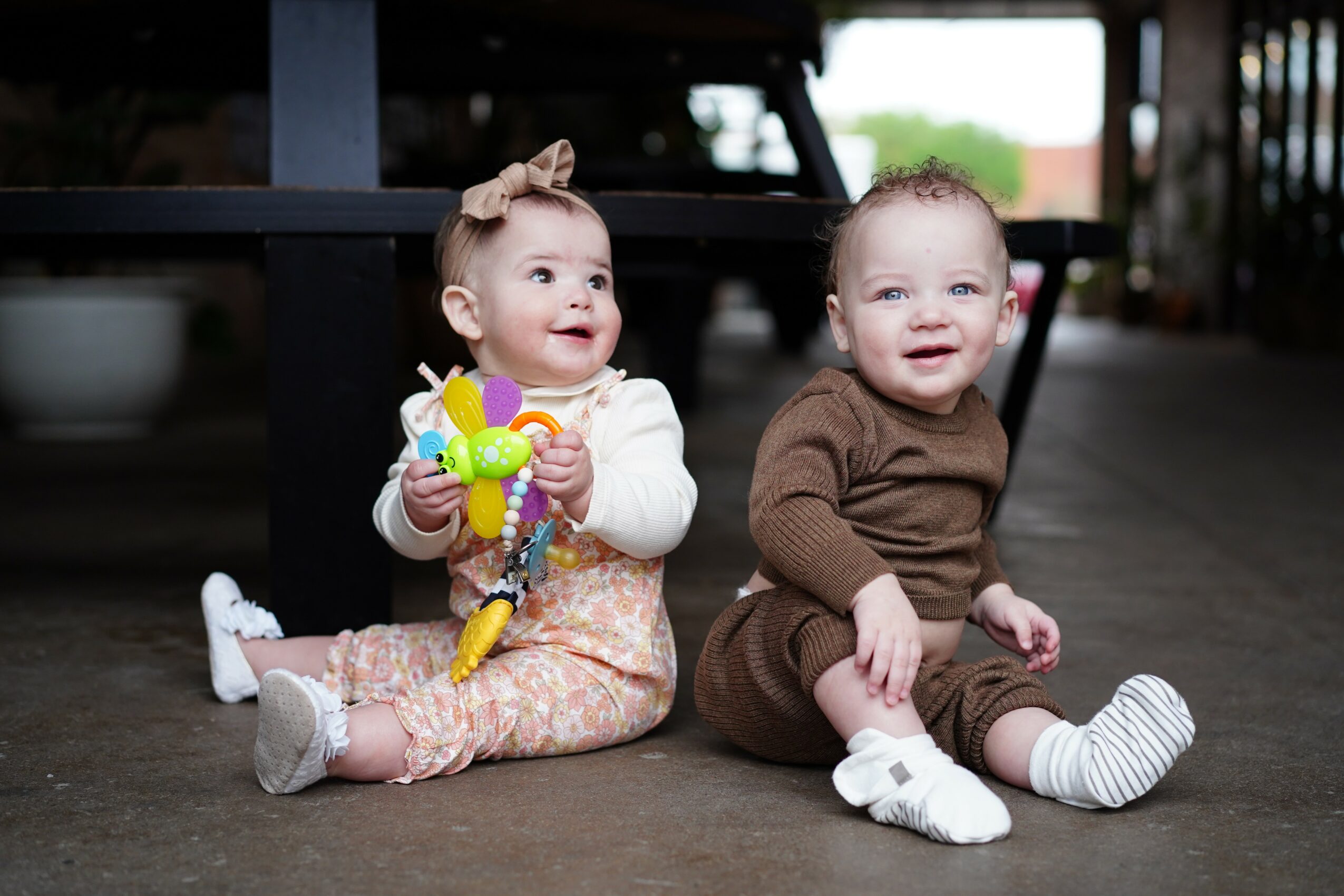 Two young children sitting on the floor, one holding a colorful toy, smiling and looking to the side.