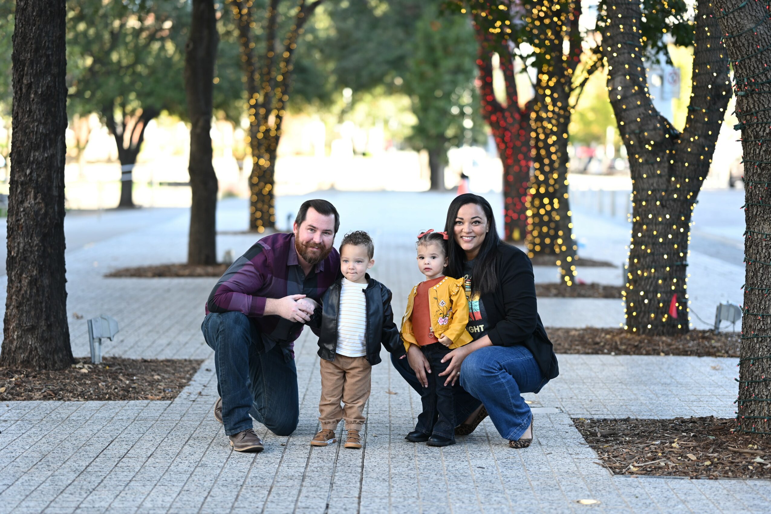 Family of four outdoors in a park with decorated trees, smiling, standing on a paved path.