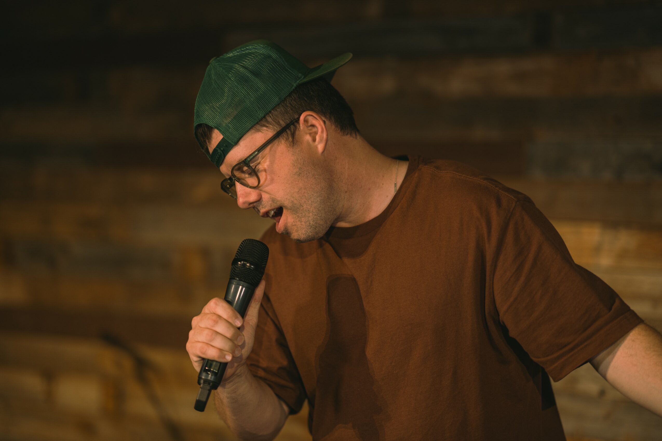 Man wearing glasses and a green cap holding a microphone, speaking or singing, with a wooden background.