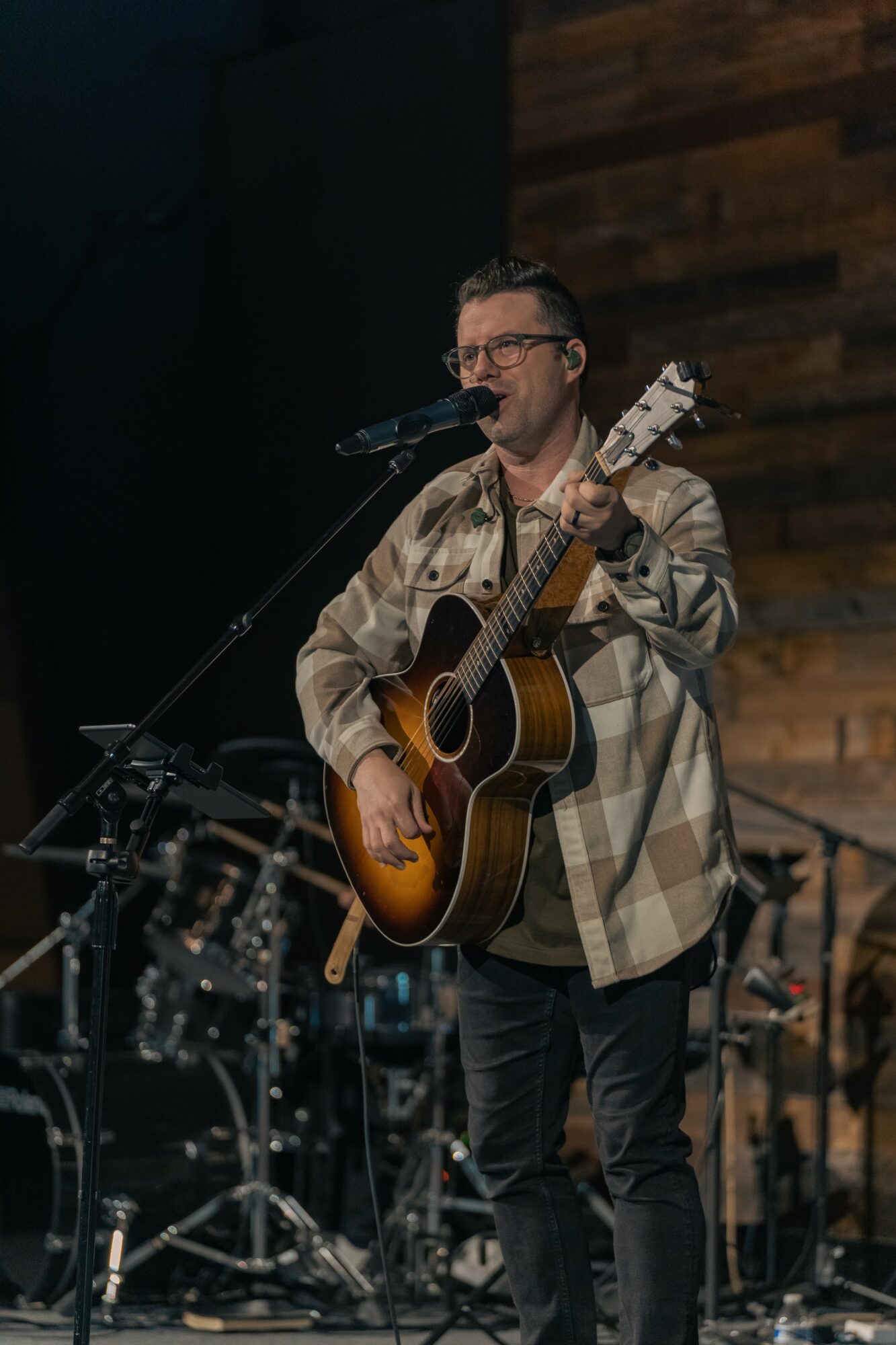 Man singing and playing guitar on stage with drums and wooden wall background.