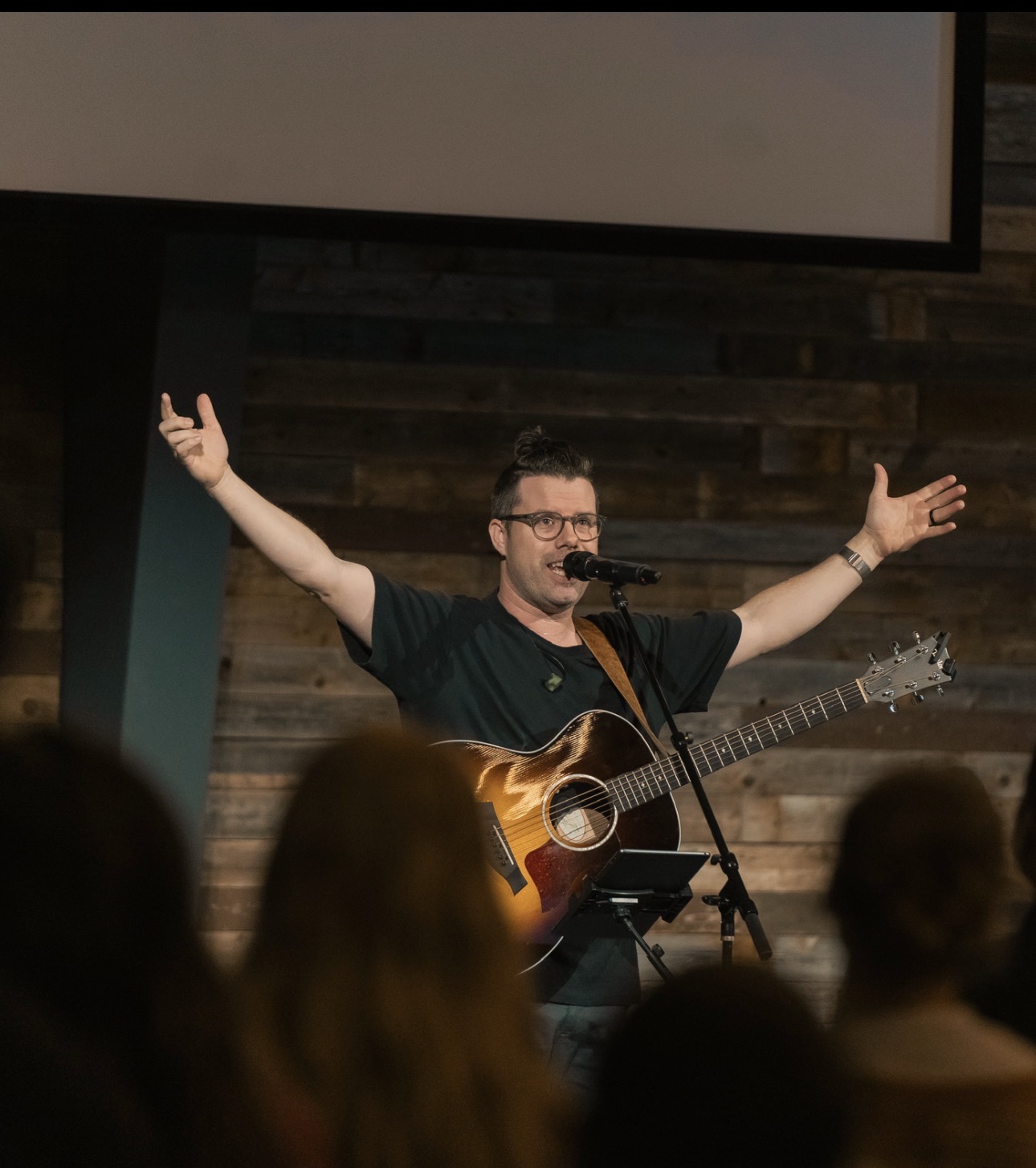 Man with glasses singing into a microphone, playing an acoustic guitar, with arms outstretched on stage in front of an audience.