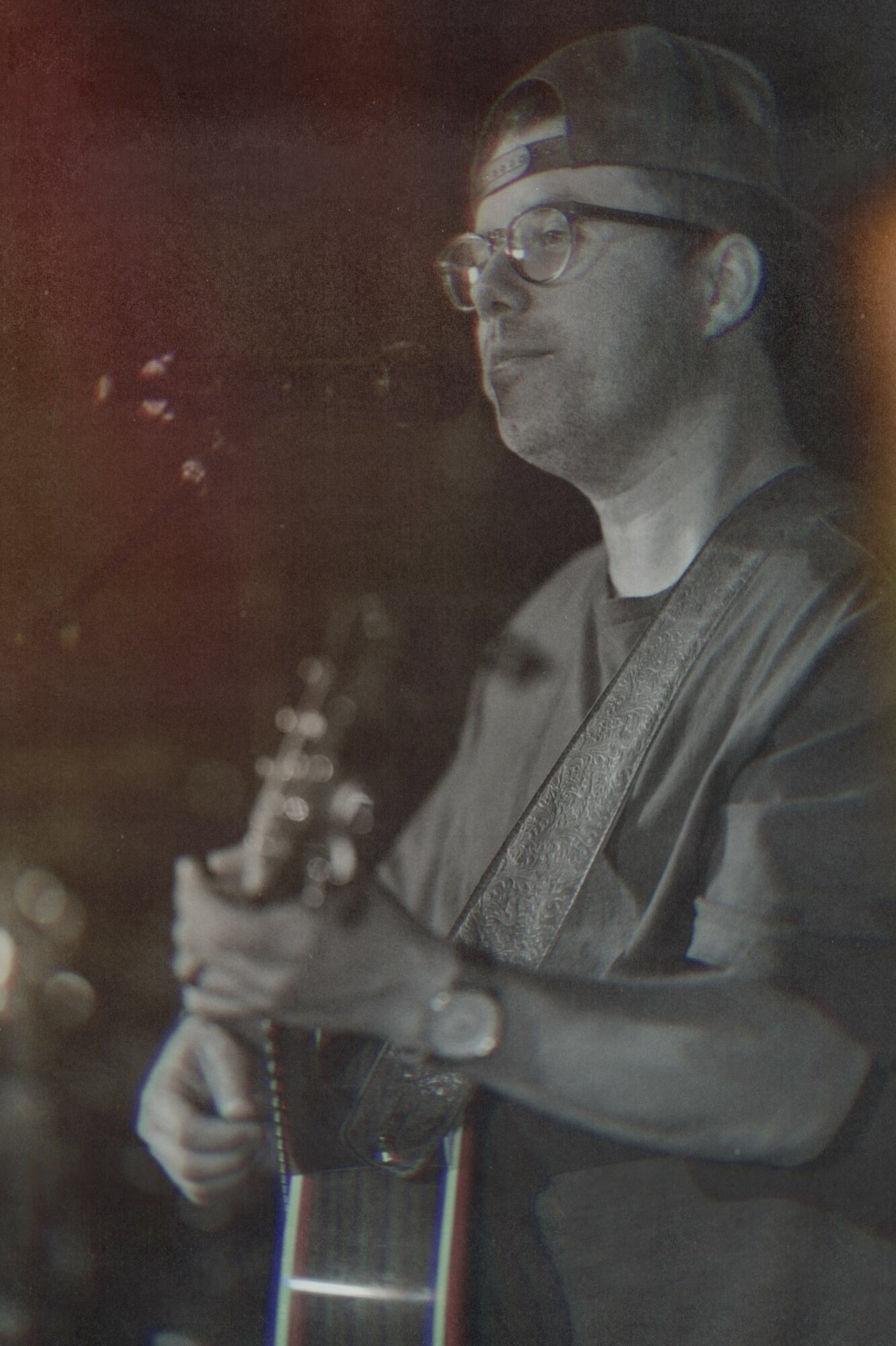 Person wearing glasses and a cap playing guitar, looking to the side, in a dark setting.