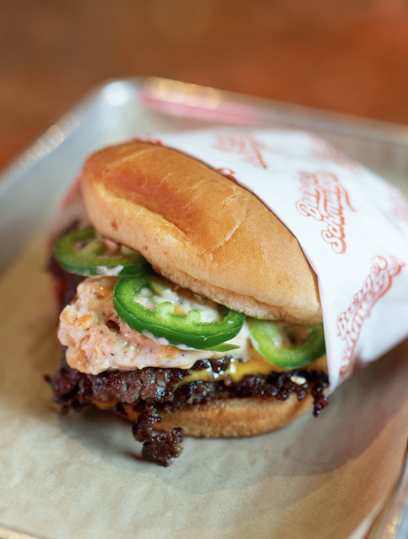 A burger with a bun, jalapeño slices, a patty, and sauce, partially wrapped in paper, on a tray.