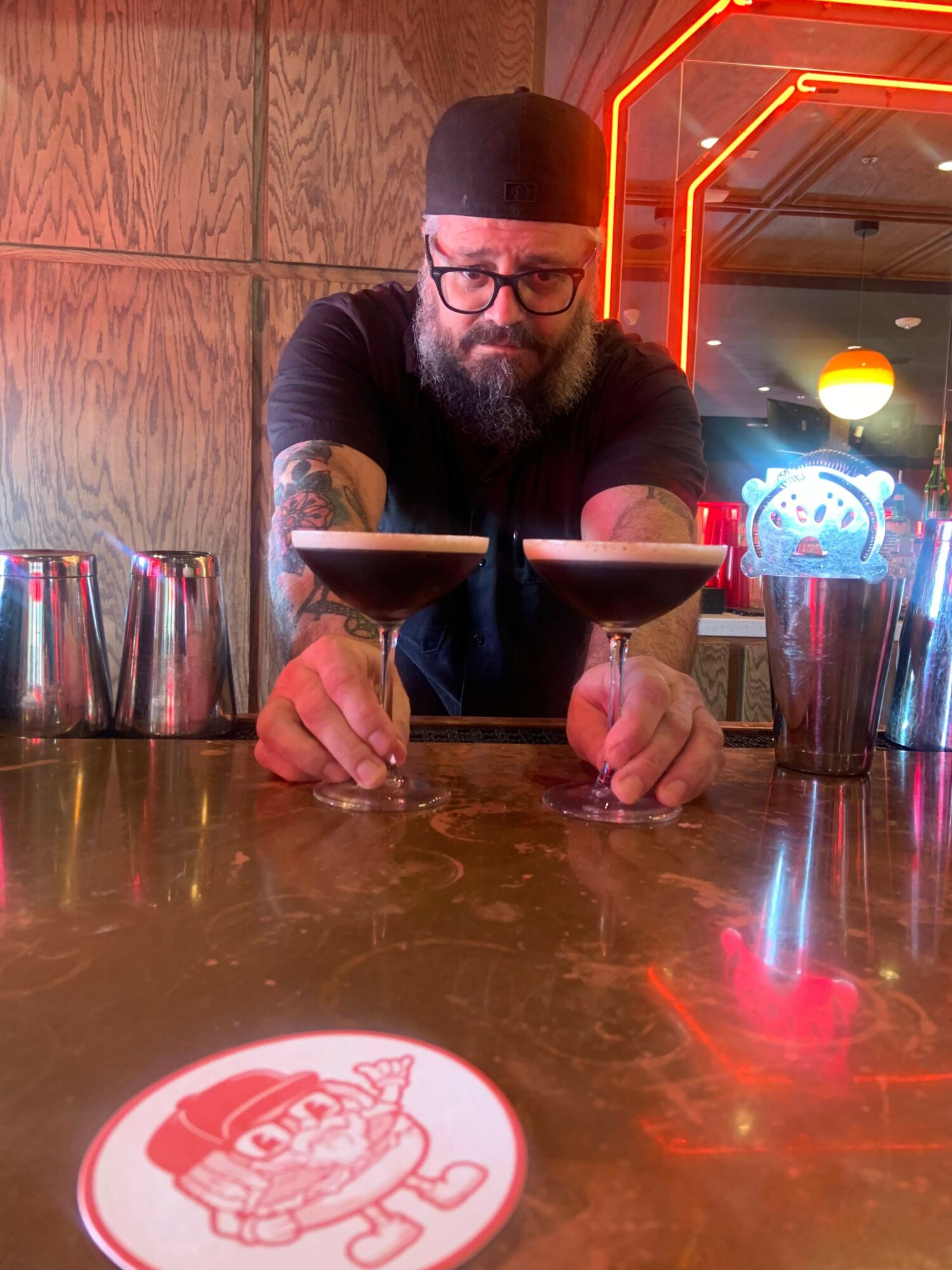 Person with glasses and beard sitting at a bar, writing on a coaster, with neon lights and a wooden wall behind.