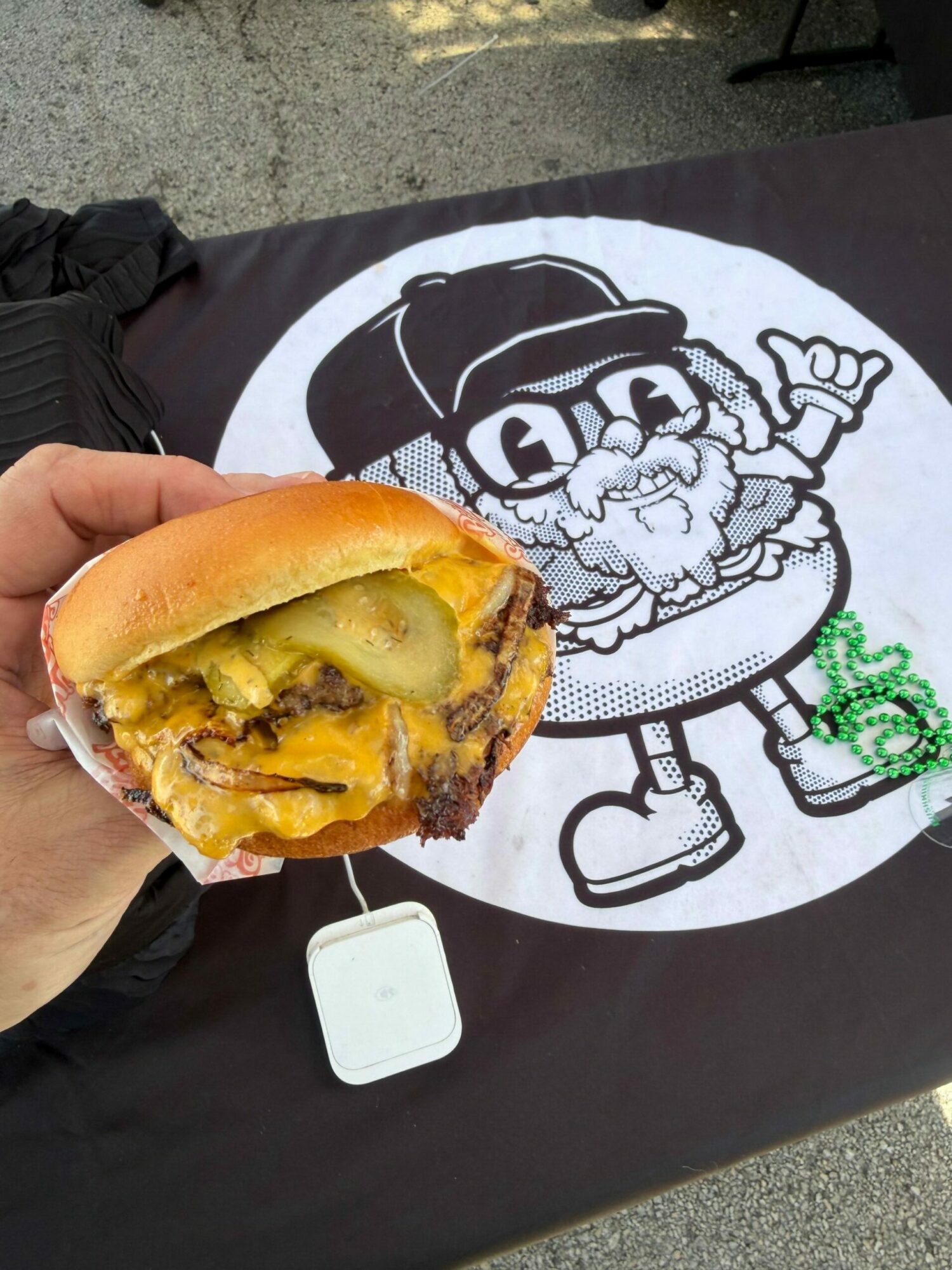 Person holding a hot dog with mustard, pickles, and meat, above a black table with a cartoon drawing of a character. Word 'Dino' visible.