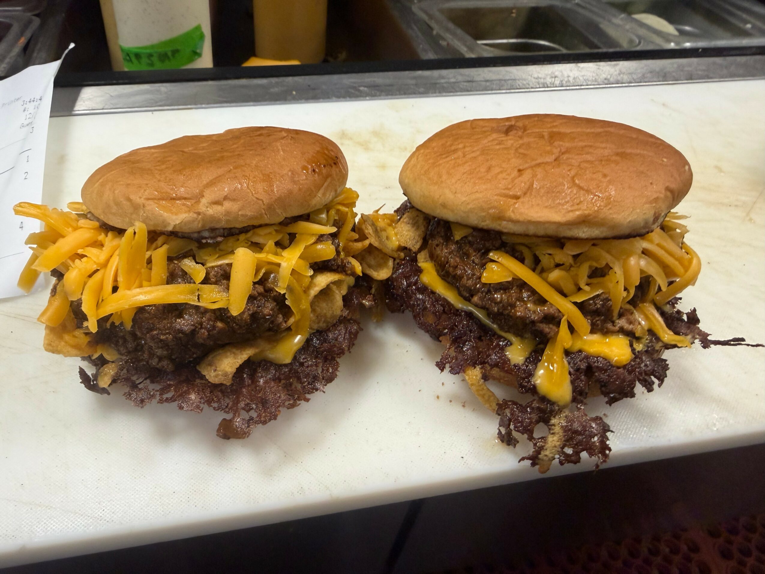 Two cheeseburgers with beef patties, shredded cheese, and buns on a white surface.