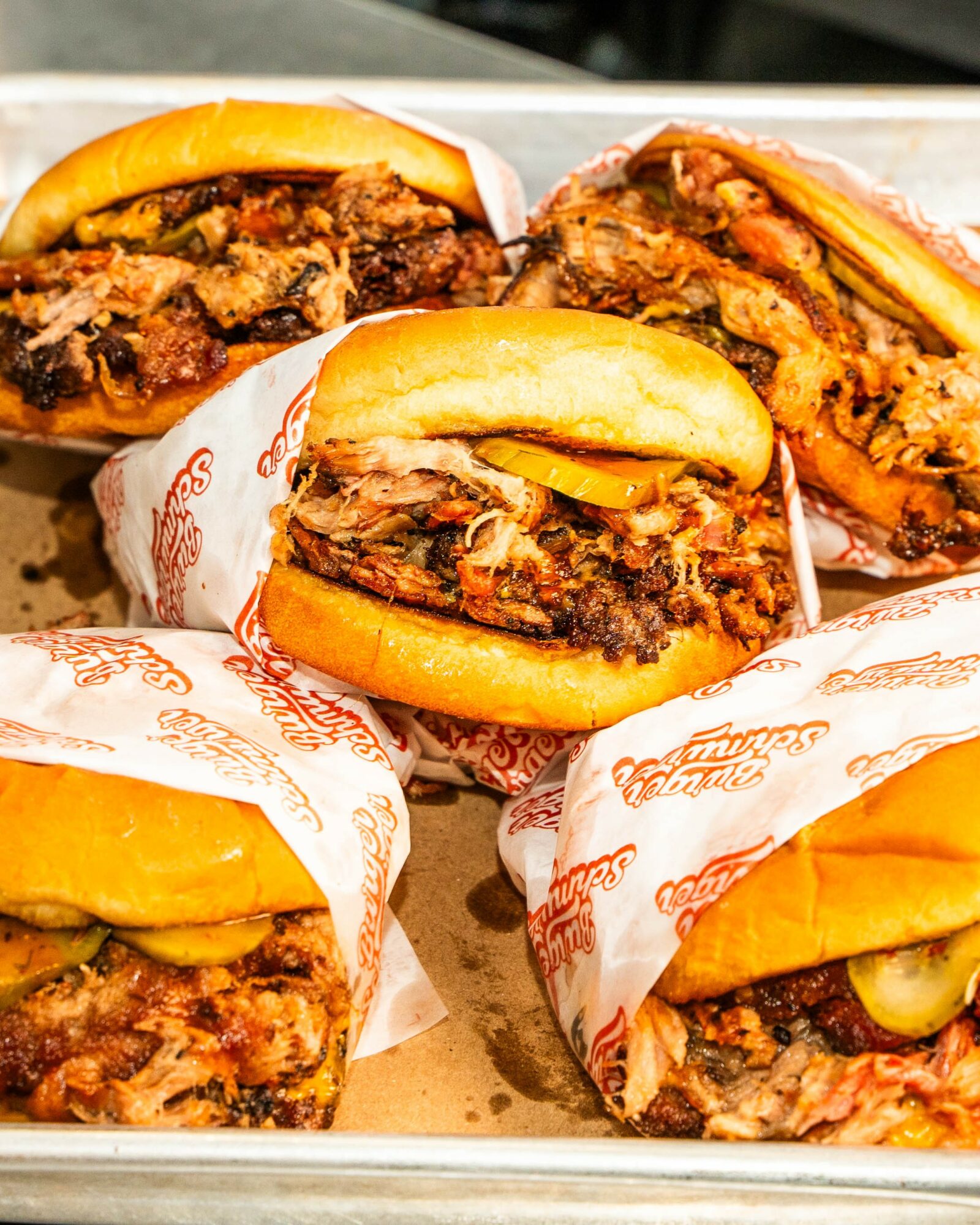 Four cheeseburgers with beef patties, pickles, and buns, wrapped in branded paper, arranged on a tray.