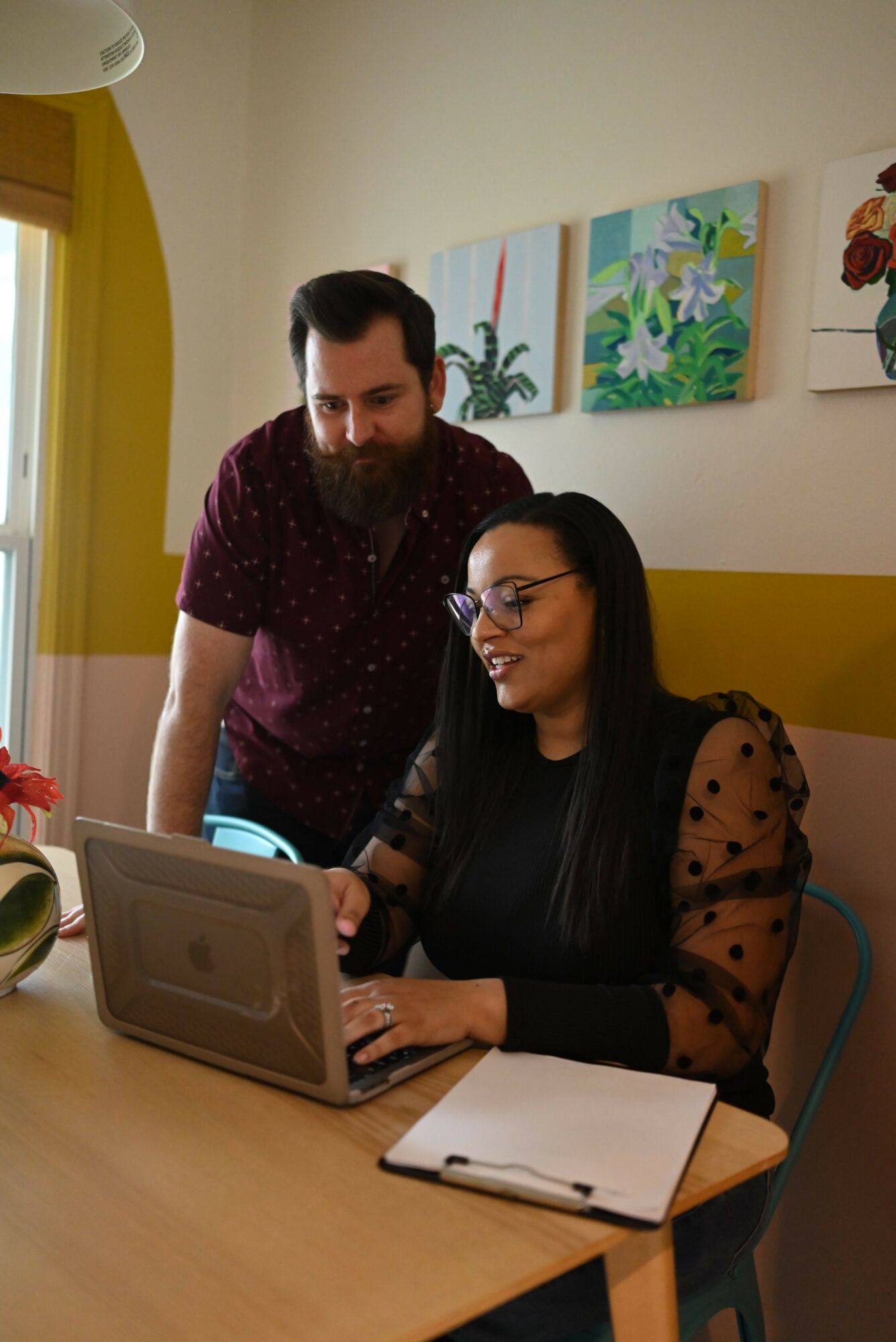 Two women sit at a table, one using a laptop, in a room with colorful artwork on the wall.