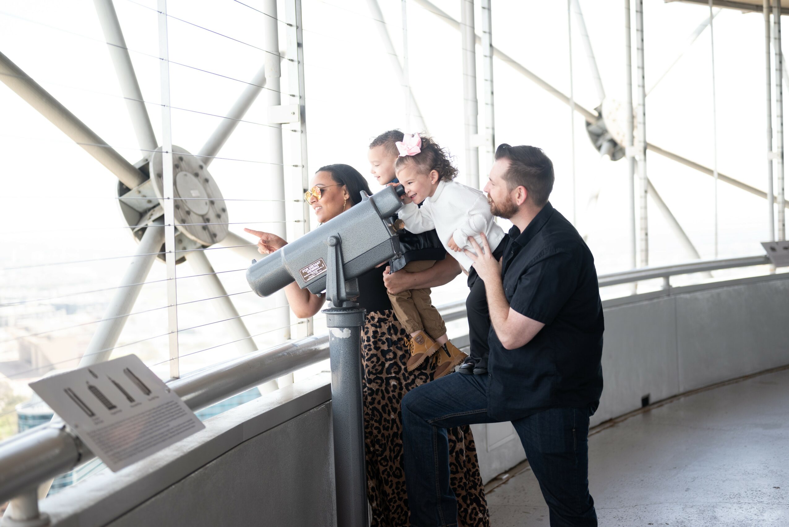 Three people, including a child, look through a telescope at a museum or observatory. They are near a large window with metal framework.