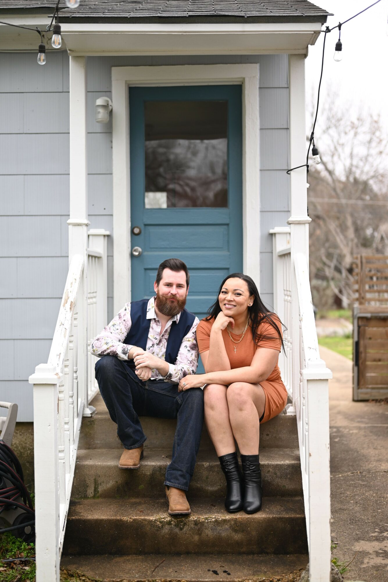 Two women sitting on front porch steps, one with long hair and the other with short hair, in front of a blue door.