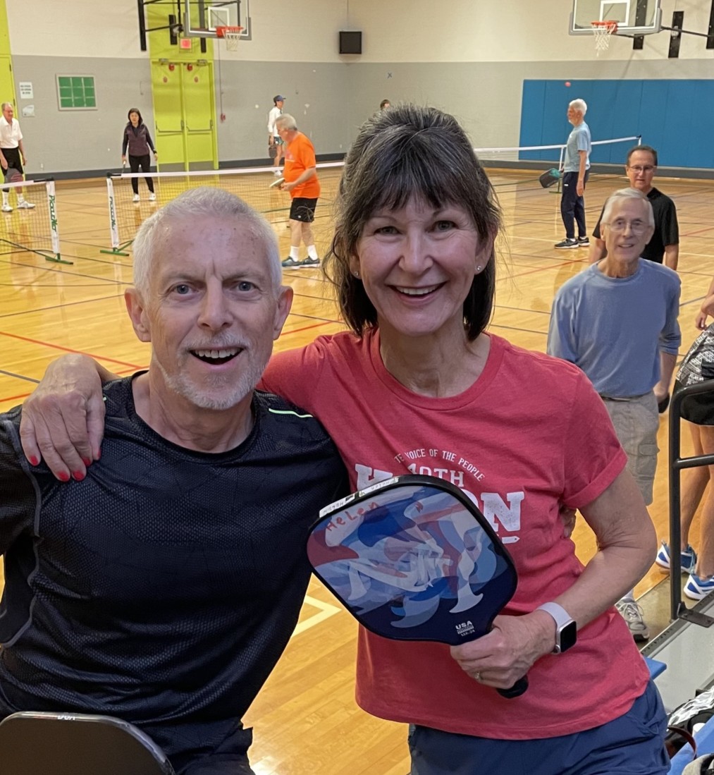 Two smiling people, a man and a woman, standing in a gymnasium, holding a paddle, with others playing in the background.