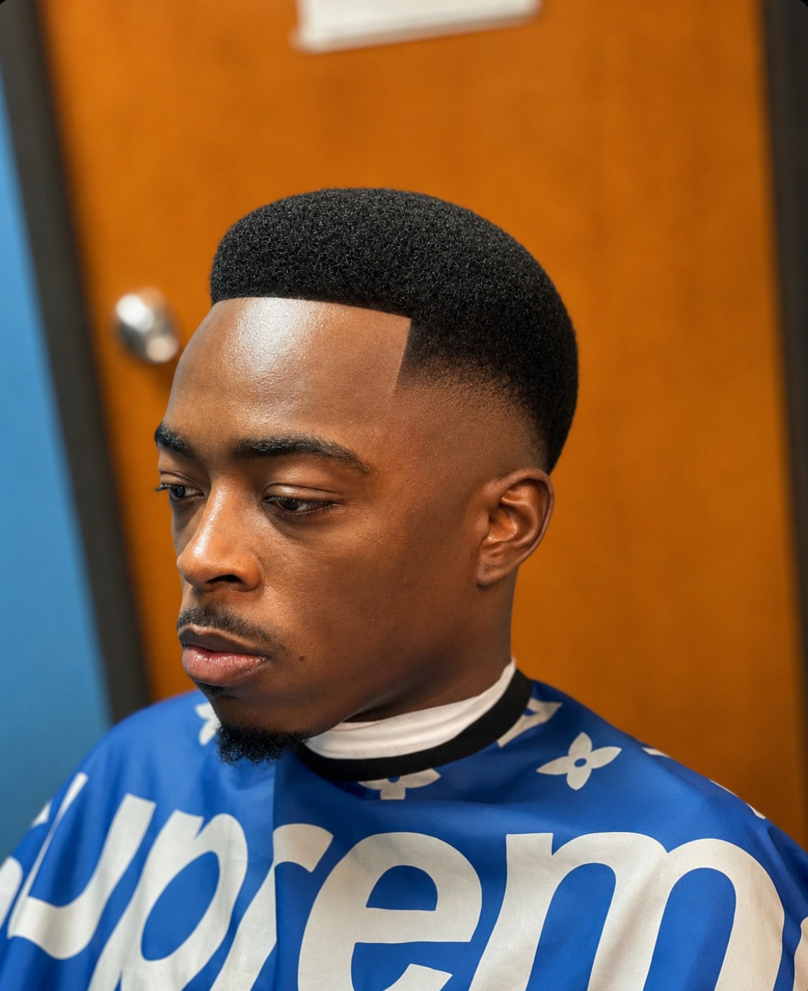 Young man with a high-top fade hairstyle, wearing a blue and white patterned shirt, looking to the side.