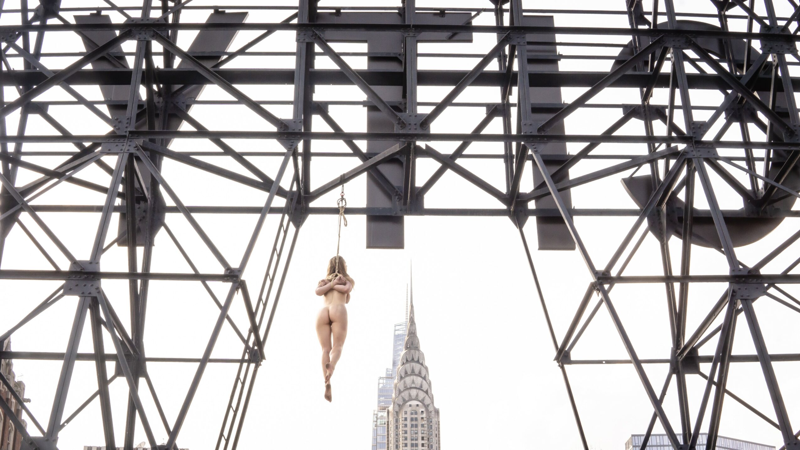 A person hanging from a crane with a tall building in the background, under a large metal structure.