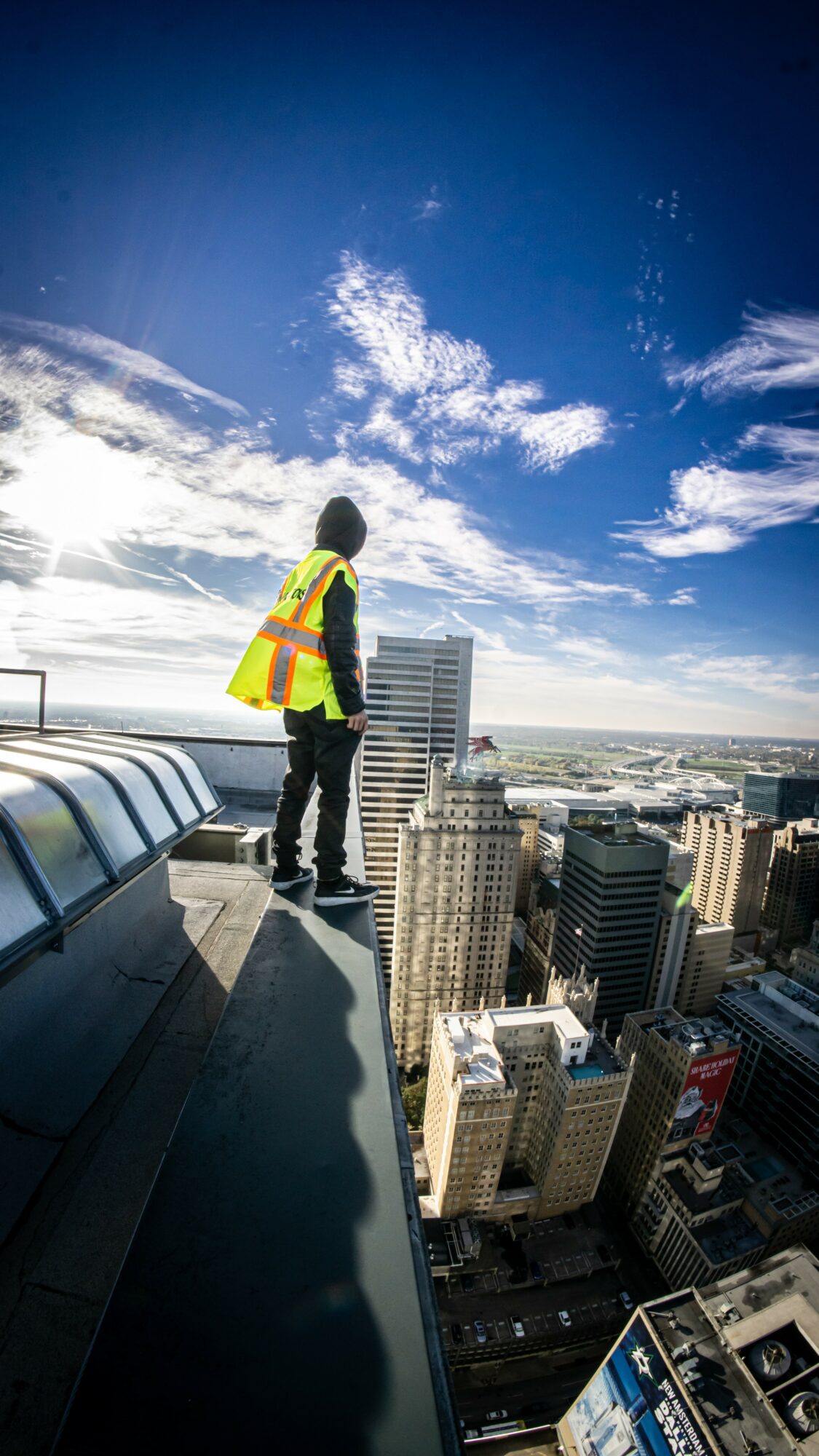 Person standing on rooftop edge overlooking city skyline with tall buildings and blue sky with clouds.