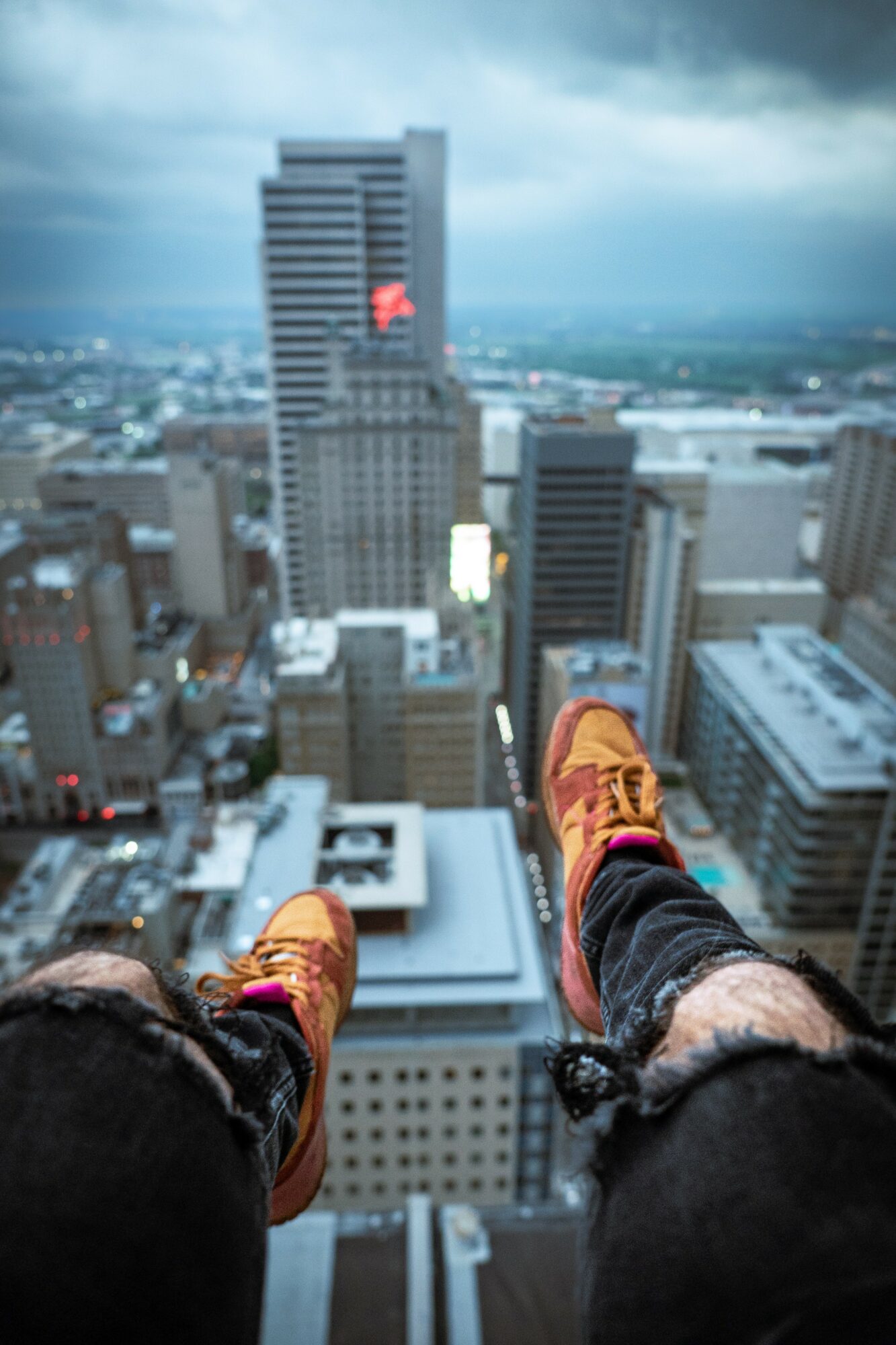 Person's legs and orange shoes dangling over city skyline with tall buildings and cloudy sky.