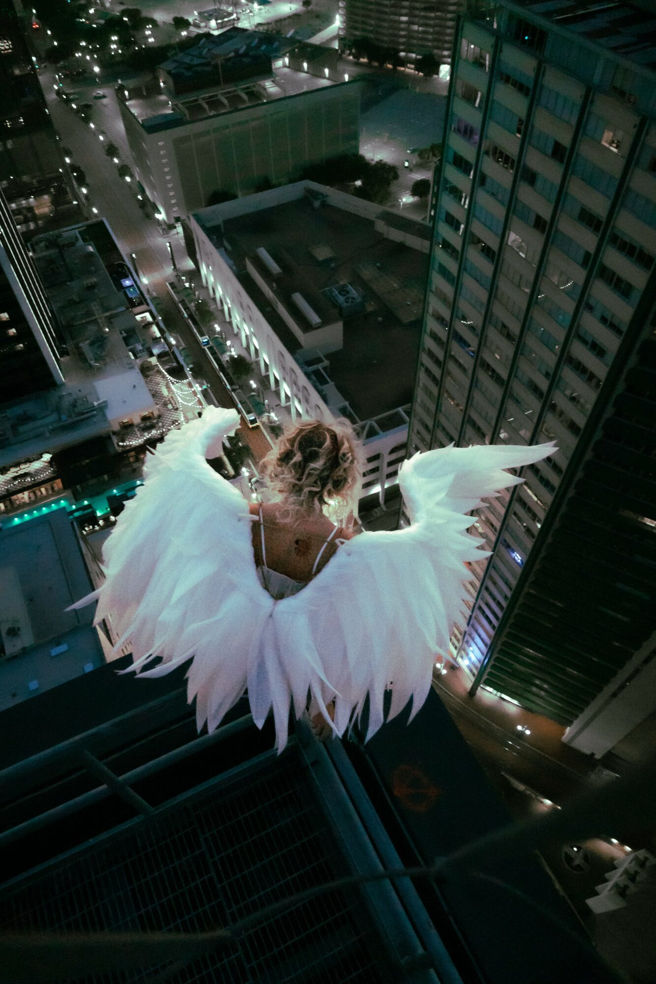 Person dressed as a swan with large white feathers sitting on a rooftop ledge at night, cityscape in background.